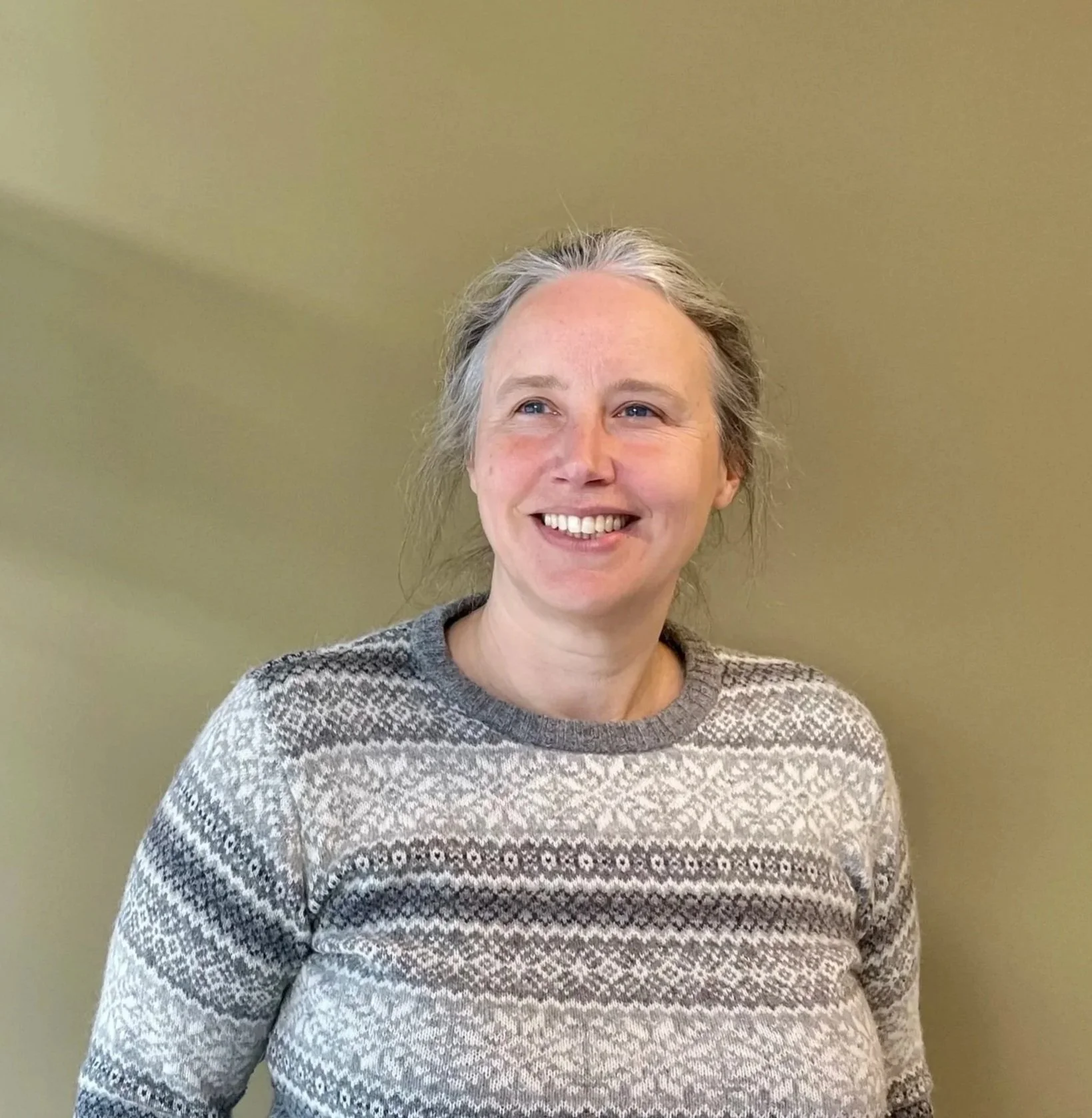 Woman with gray hair smiling in front of beige wall, wearing a gray patterned sweater.