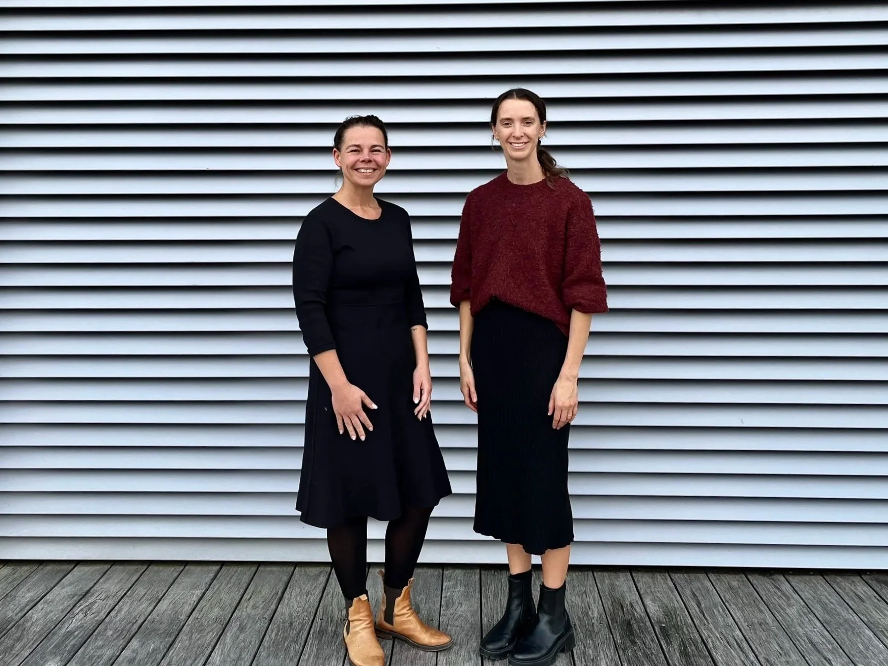 Two women smiling and standing side by side on a wooden deck in front of a gray metal horizontal siding wall. The woman on the left is wearing a black long-sleeve dress with a flared skirt and tan ankle boots. The woman on the right is wearing a maroon sweater, black skirt, and black ankle boots. Both have dark hair and appear to be outdoors.
