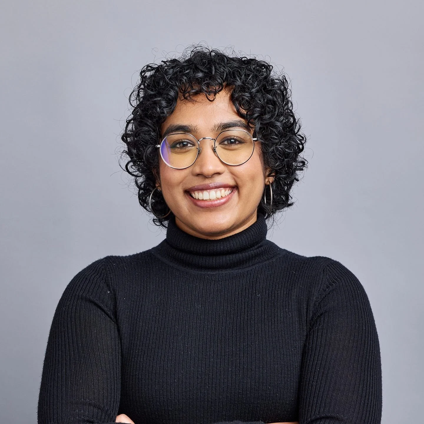 A woman with curly black hair, wearing glasses, hoop earrings, and a black turtleneck sweater, smiling against a plain gray background.