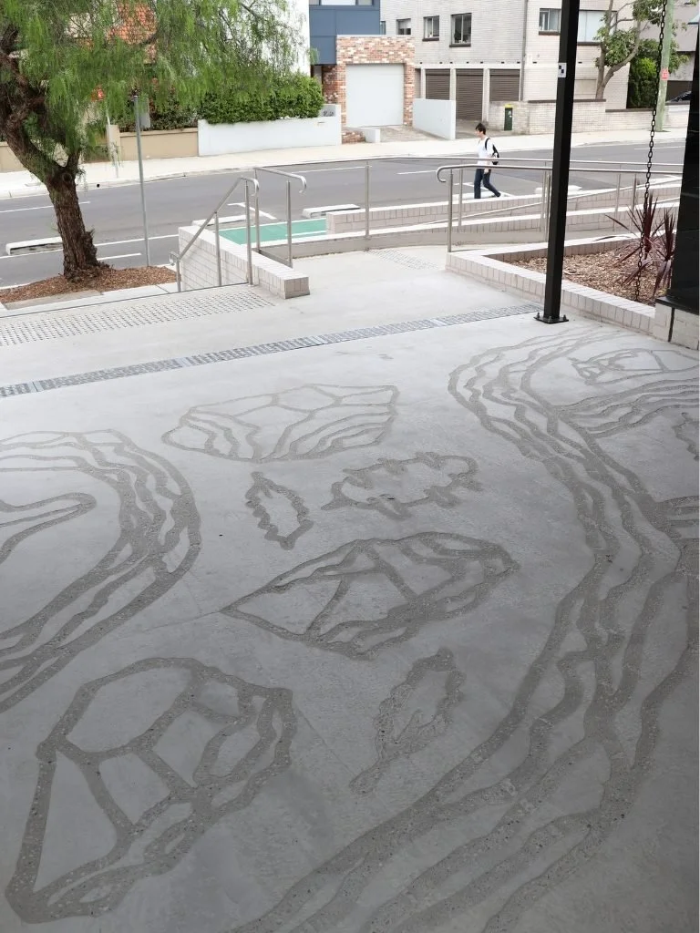 Concrete floor with abstract sand patterns resembling a landscape, trees, and water, viewed from inside a building with an open view to the street and a person walking outside.