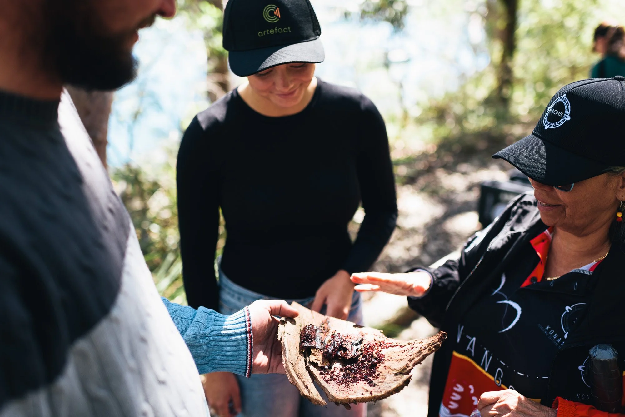 Group of people outdoors during daytime, one person holding a piece of wood with dark staining, others gathered around looking at it, trees in background.