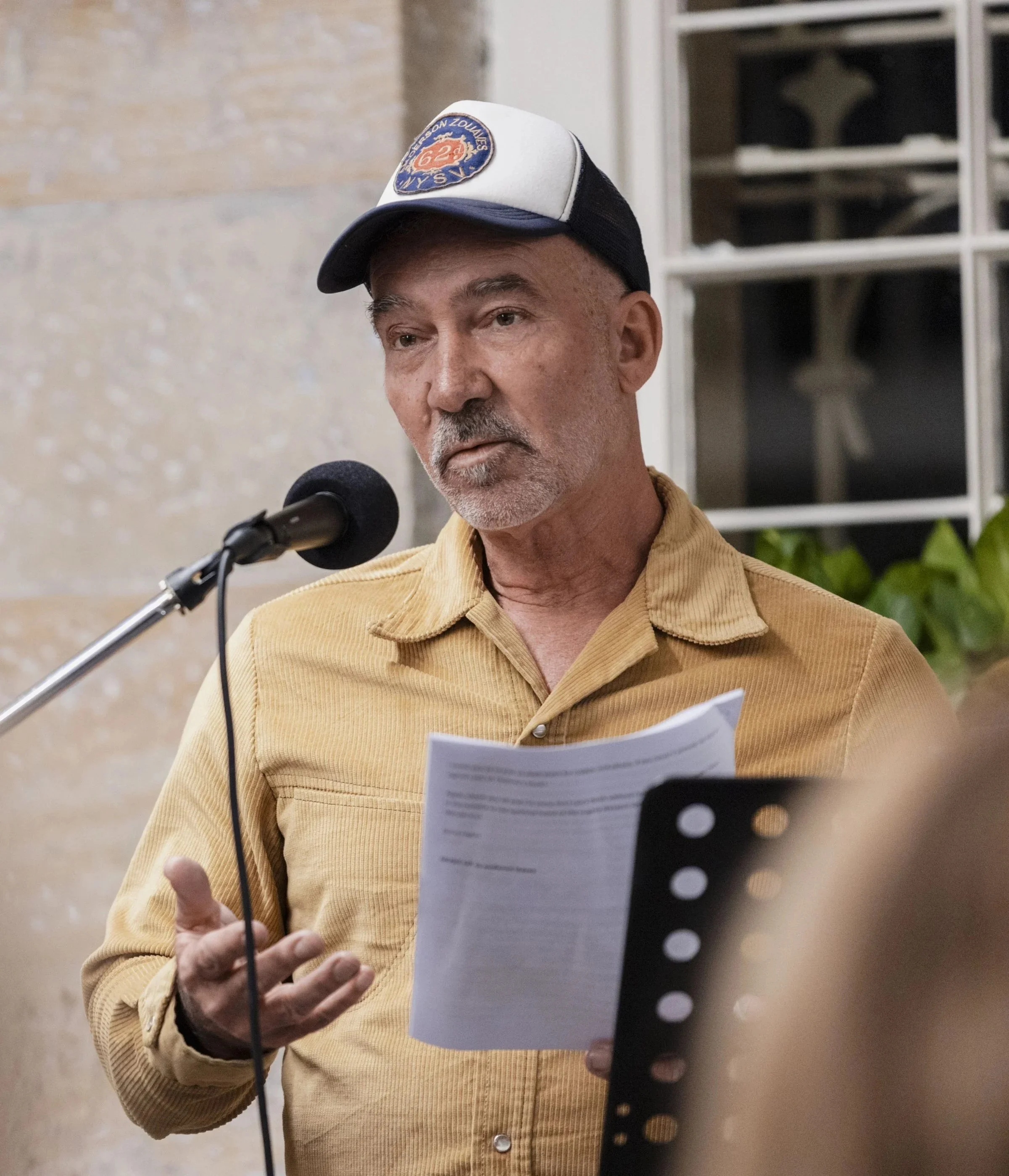 A man wearing a beige corduroy shirt and a trucker hat with a patch, speaking into a microphone and holding a piece of paper, standing indoors near a textured wall and a window with dark glass.