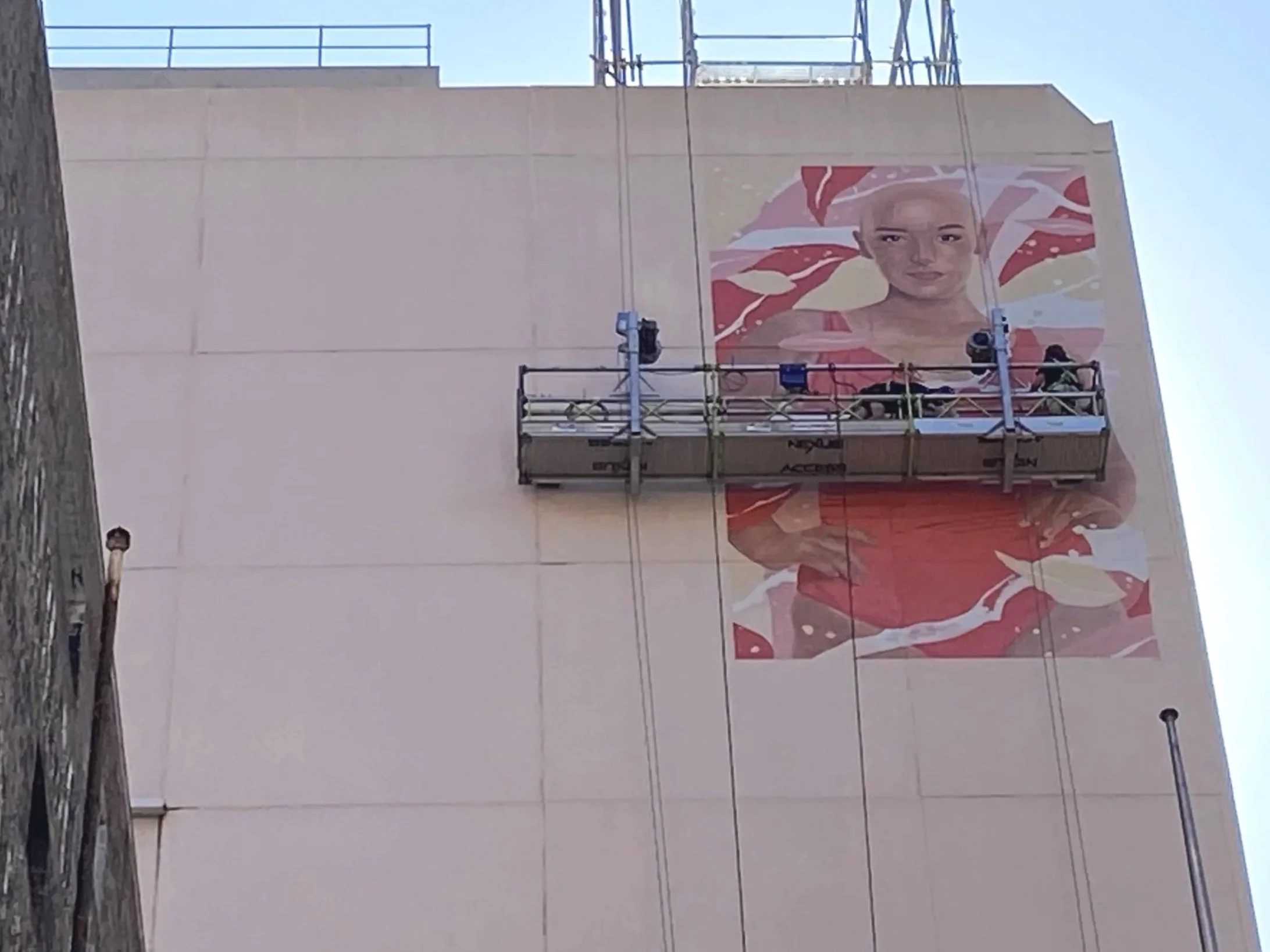 Workers on a scissor lift installing a large poster of a woman on the side of a building, with the poster featuring a woman with a headscarf and red clothing.