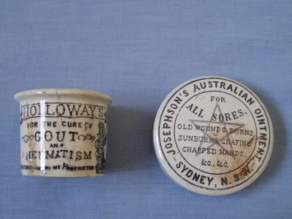 Two antique medicinal containers with printed labels related to old Australian remedies. One is a small ceramic cup with text for the cure of gout and rheumatism. The other is a round tin with a warning about sores and old wounds.