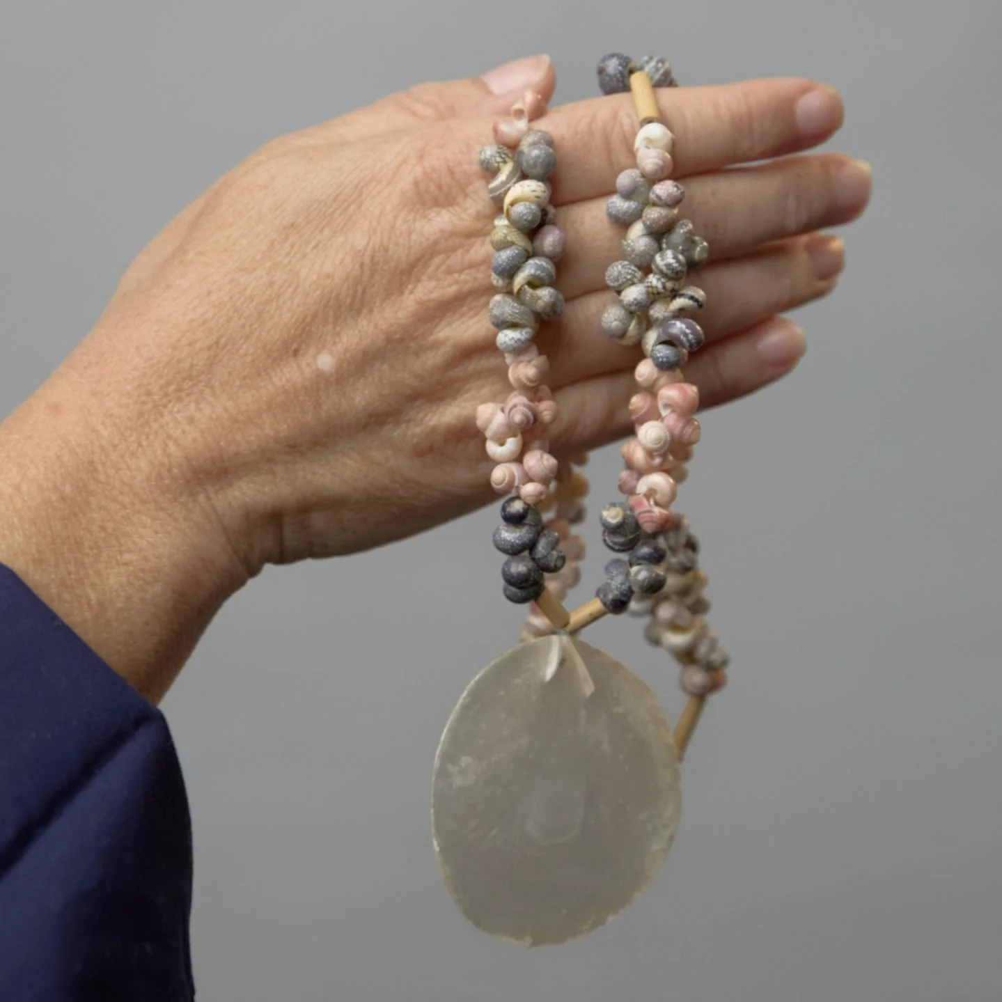 A person's hand holding a beaded necklace with shells and a large circular pendant, against a gray background.