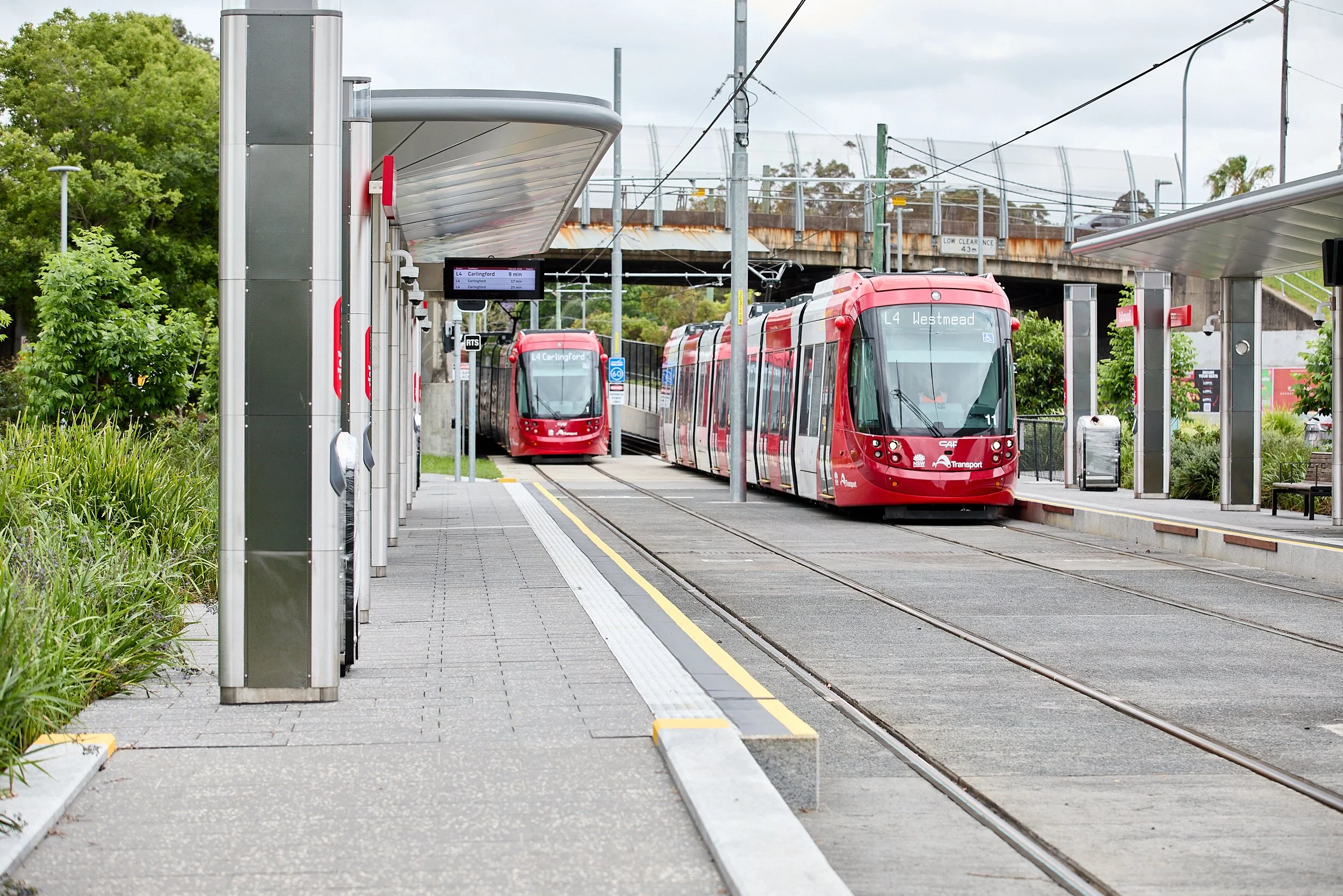 Red tram at a modern tram station with digital display and overhead electrical wires, surrounded by greenery and a railway bridge in the background.