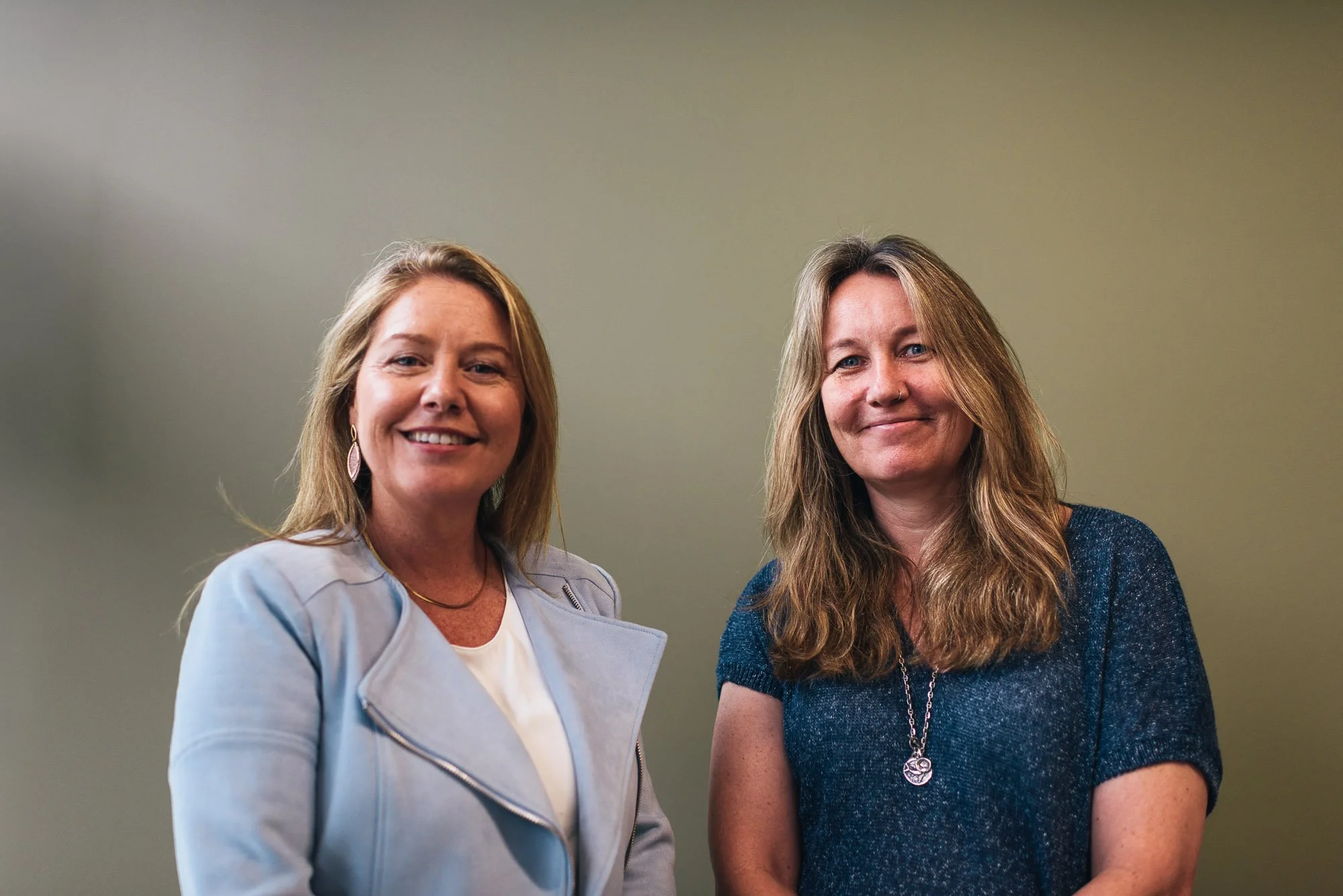 Two women standing next to each other, smiling, against a plain green wall. The woman on the left has long, light brown hair and is wearing a light-colored jacket over a white top. The woman on the right has long, wavy, light brown hair and is dressed in a dark blue top with a necklace.