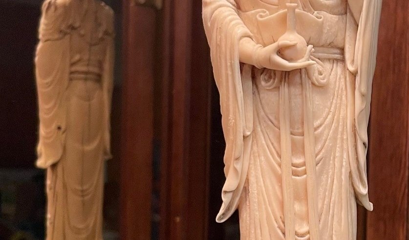 Close-up of an ivory-colored carved figurine of a person holding a vessel, with a background of another similar figure and wooden surfaces.