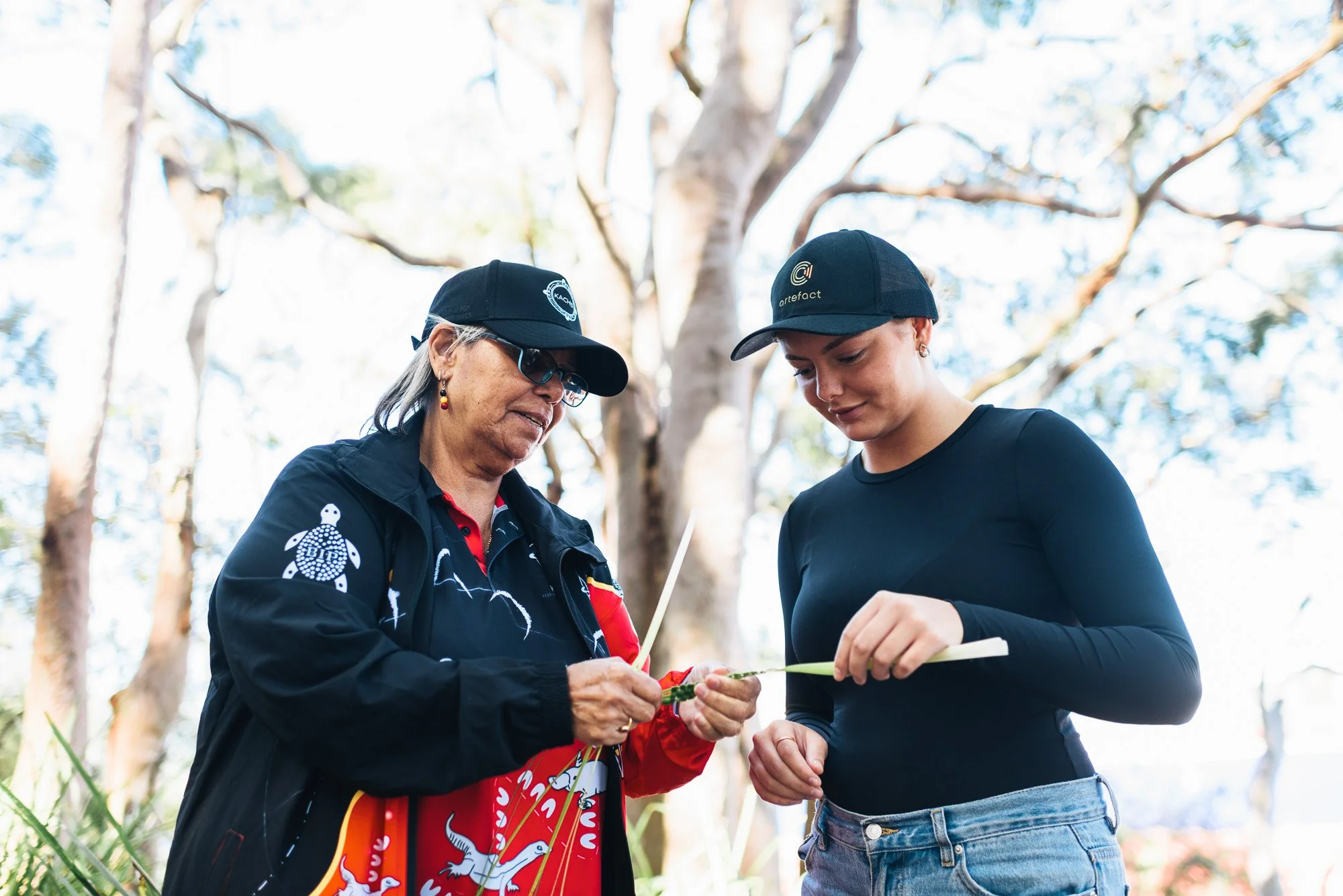 Two women, one older and one younger, stand outdoors under a tree, examining a small plant together. Both wear black caps and casual clothing, with the older woman wearing a patterned jacket and the younger in a black shirt. The background shows tree trunks and branches in a bright, natural setting.