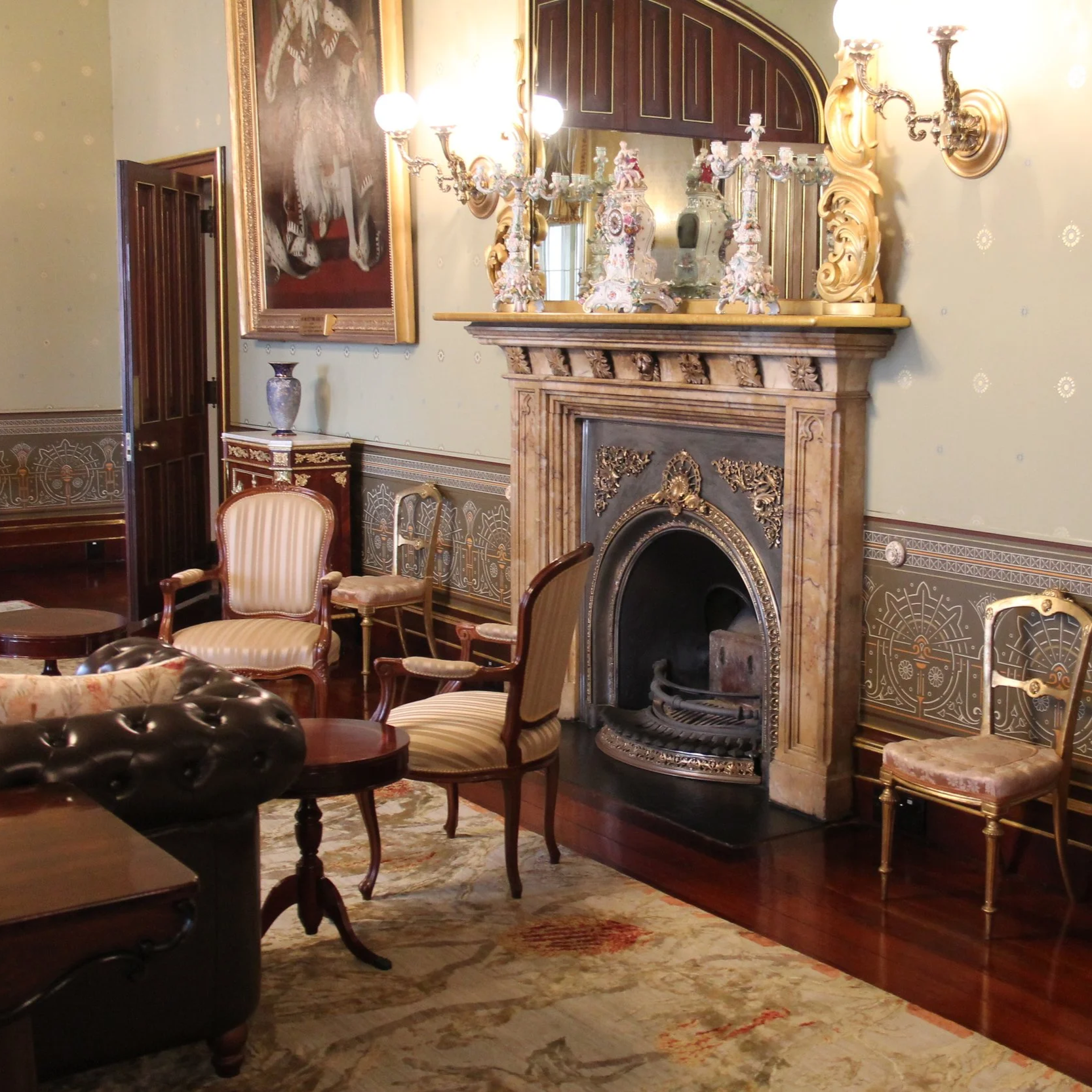 An elegant room with a marble fireplace, ornate mirror, antique chairs, a large portrait painting, and decorative wall sconces. The room features traditional wood and gold accents.