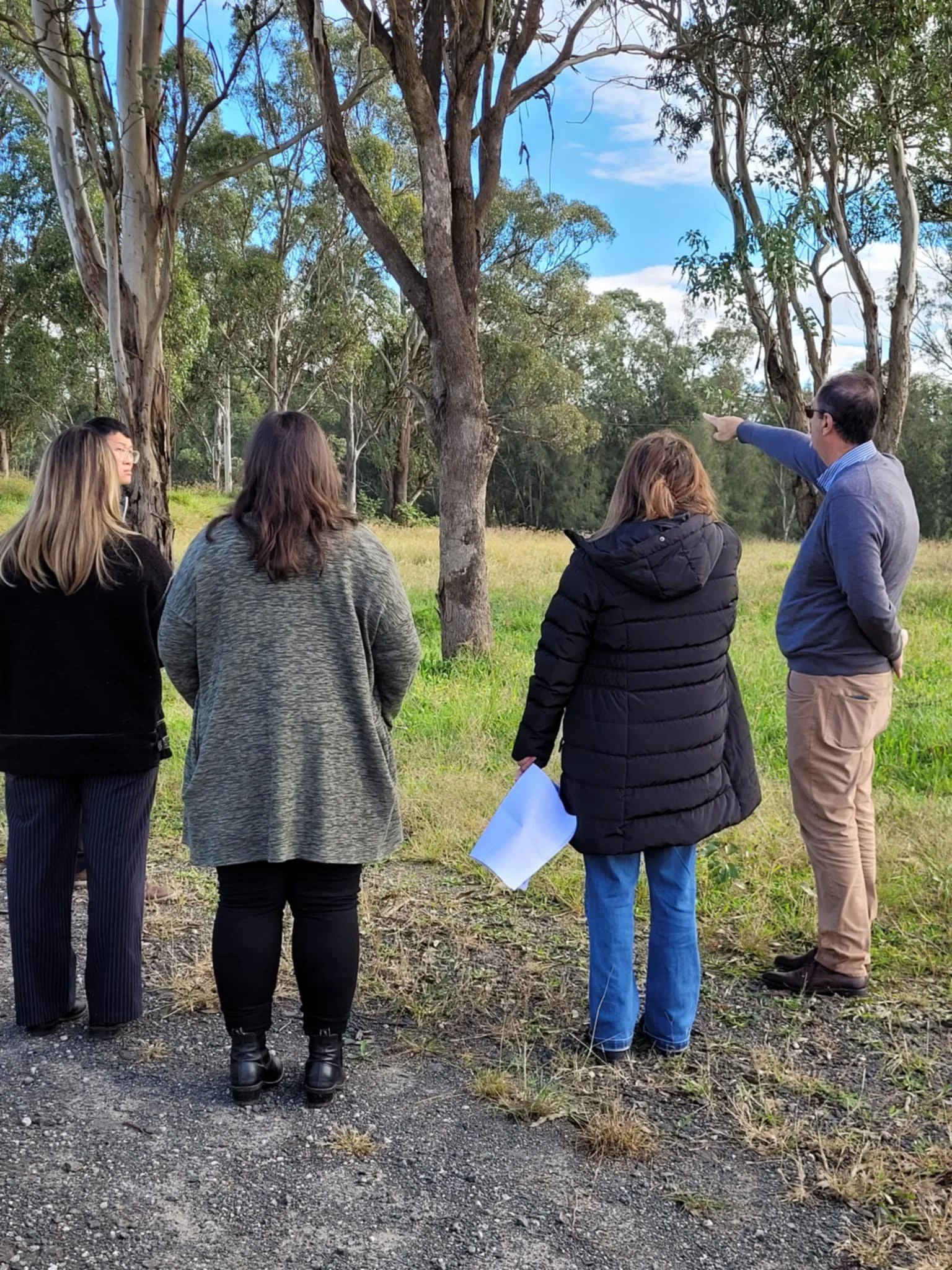 Group of five people, including three women and two men, standing outdoors near trees, listening to a man who is pointing towards the trees.
