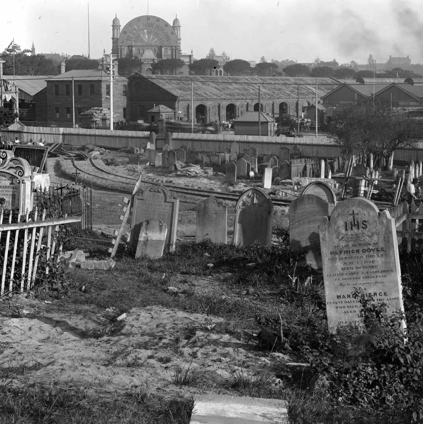 Black and white photograph of a cemetery with headstones and crosses in the foreground. A curving railroad track runs through the cemetery. In the background, there are buildings, a fence, and a large amusement park wheel or Ferris wheel with a decorative design, and possibly a roller coaster. The scene appears to be from an earlier era, possibly the early 20th century.