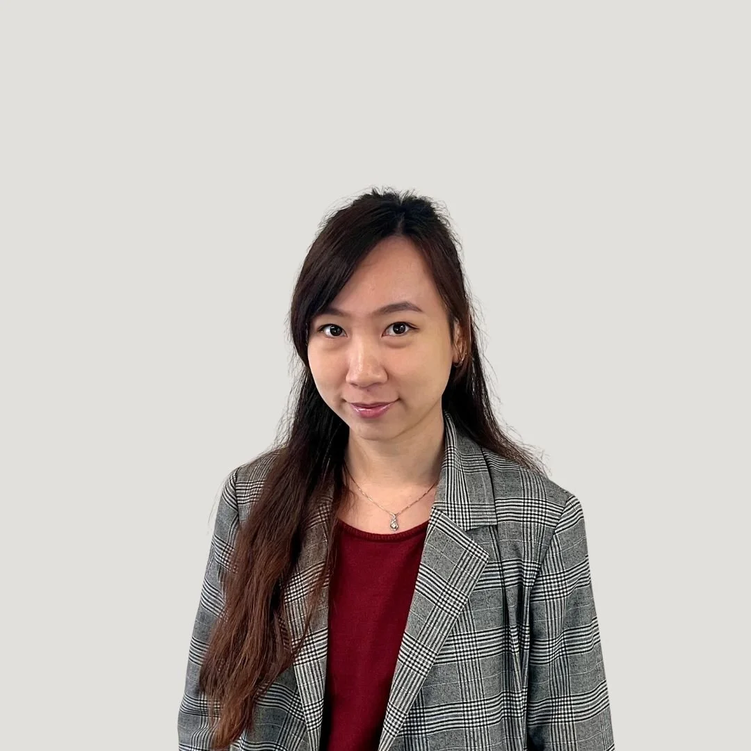 A young woman with long dark hair, wearing a checkered blazer over a red top, standing against a plain light-colored background.