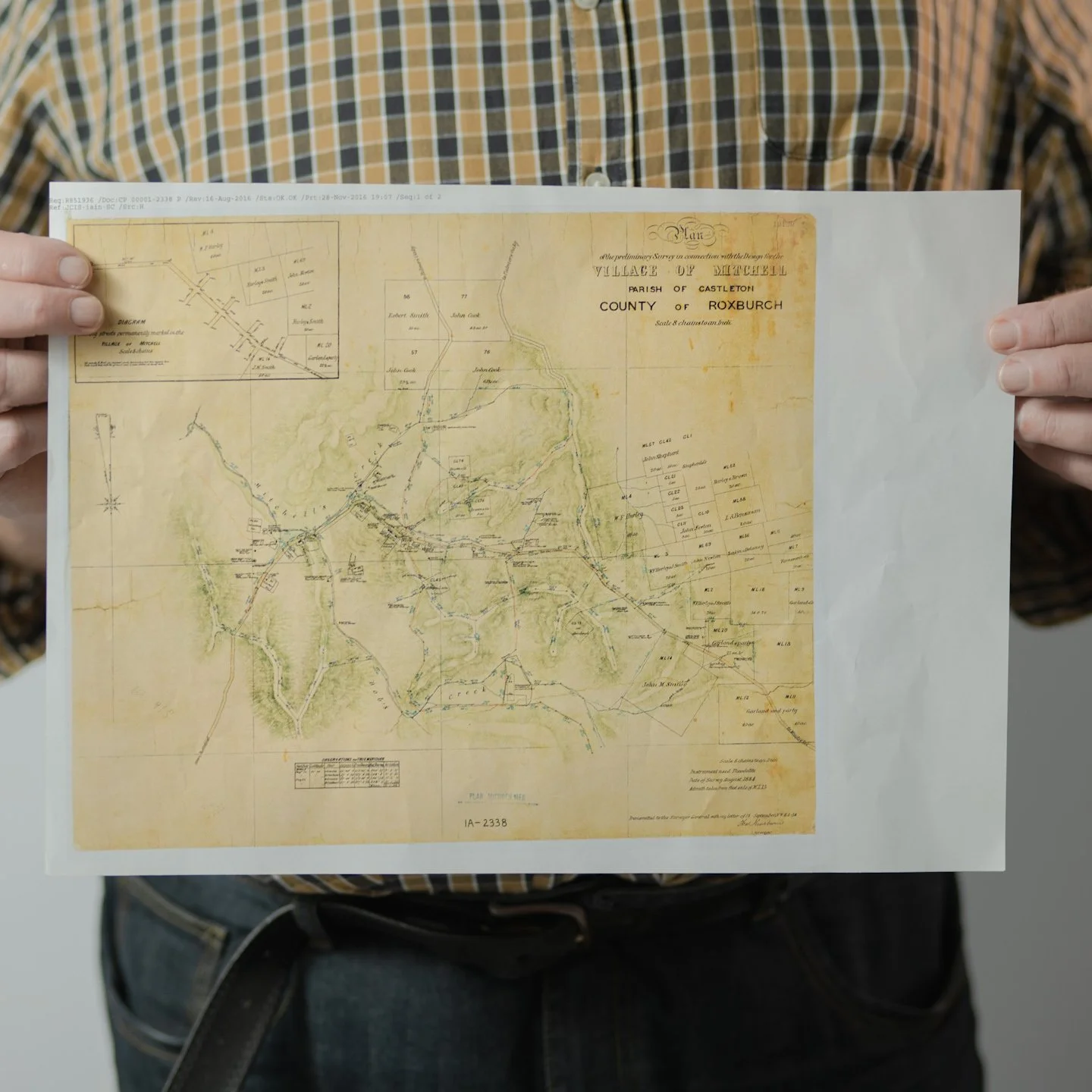 Person holding a detailed historic map of the Village of Mitchell, Parish of Castletow, County of Roxburgh, with a plaid shirt visible in the background