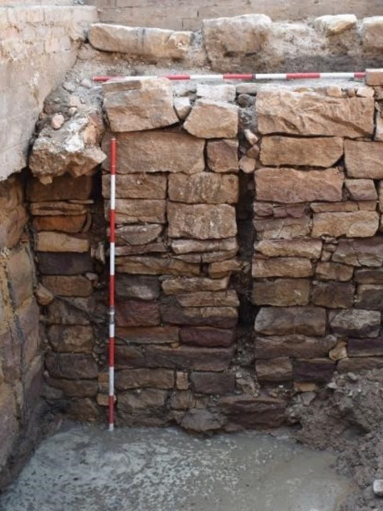 An archaeological excavation site showing a cross-section of a stone wall with a measuring rod for scale.