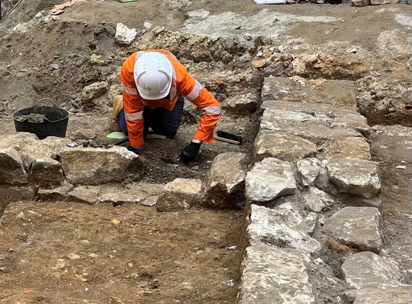 A construction worker wearing an orange safety jacket, gloves, and a white helmet is kneeling on the ground, working on a stone and dirt excavation site. There is a black bucket nearby and historic stonework is visible.