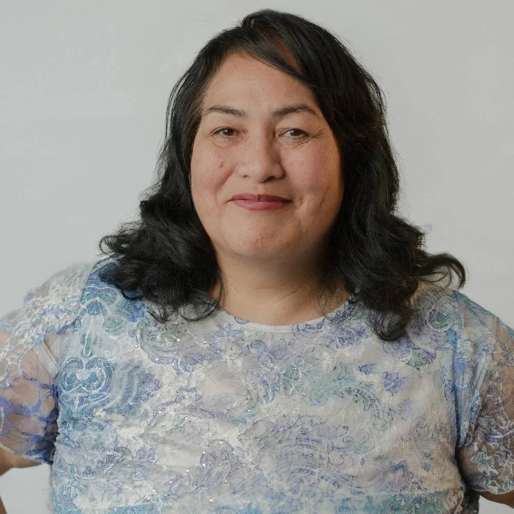 A woman with shoulder-length black hair, smiling, wearing a light-colored, floral-patterned dress with lace and glitter details, standing against a plain gray background.