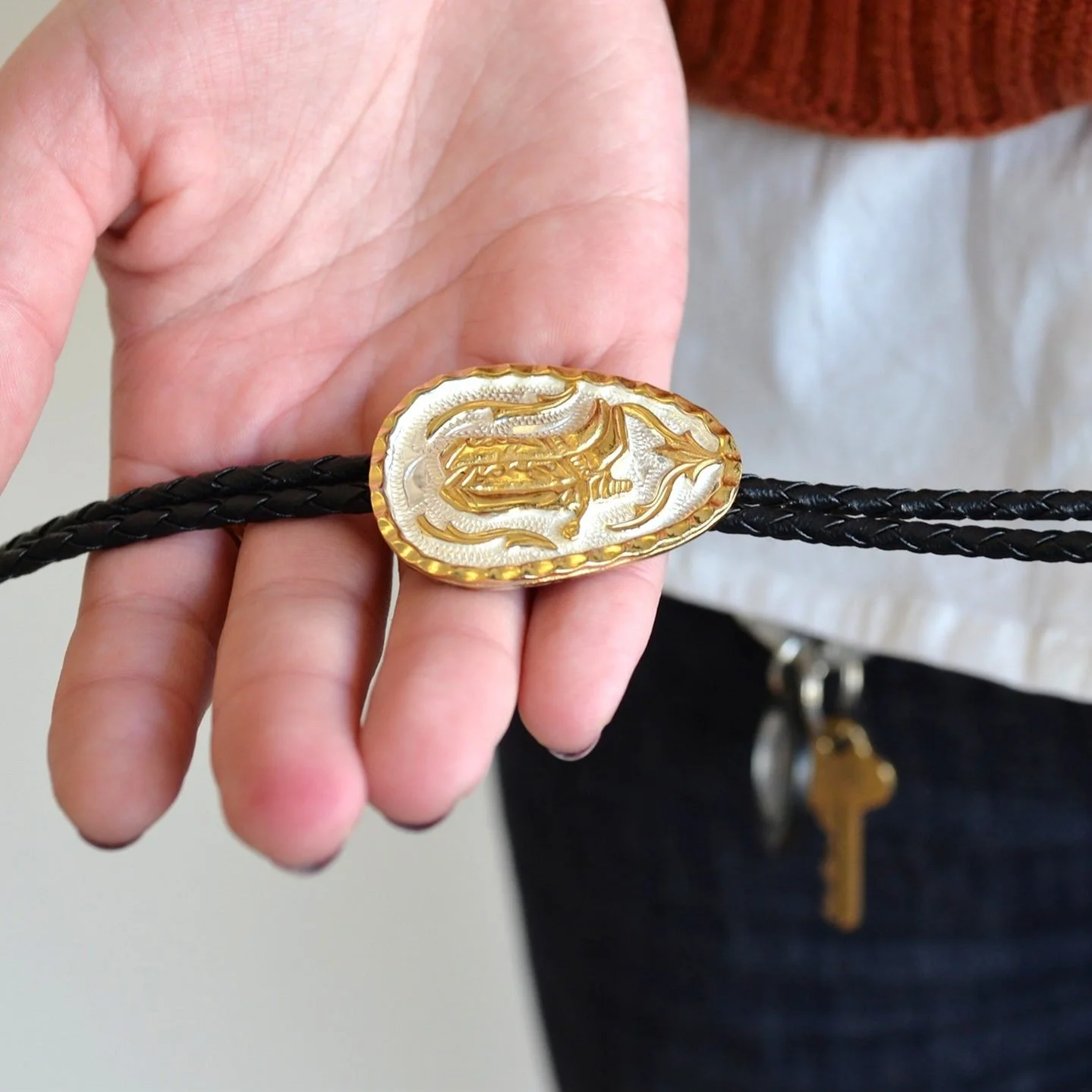 Person holding a black braided leather bracelet with an ornate gold and silver clasp featuring a curled design.