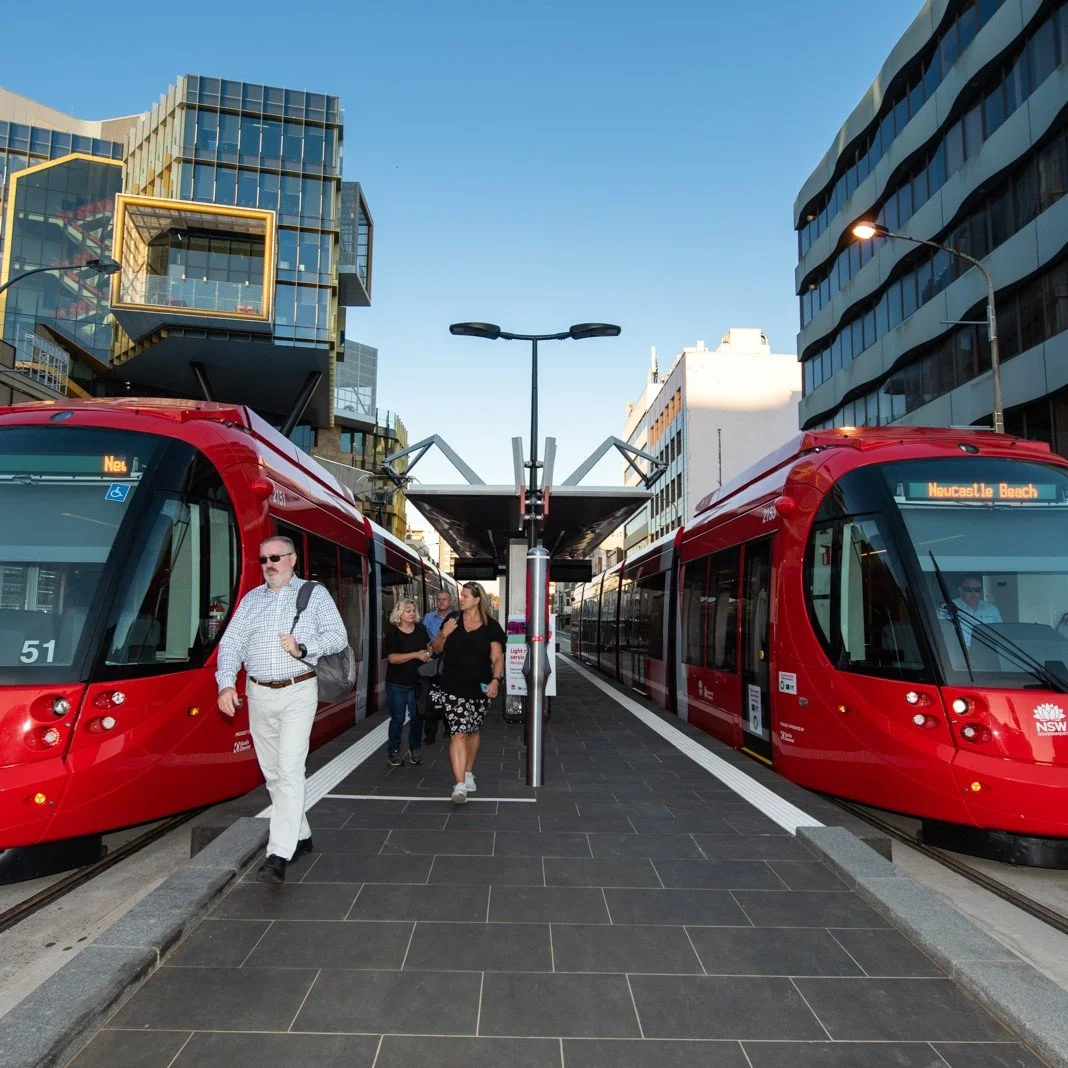 Two red trains at a modern urban train station with passengers walking on the platform, surrounded by tall buildings under a clear blue sky.