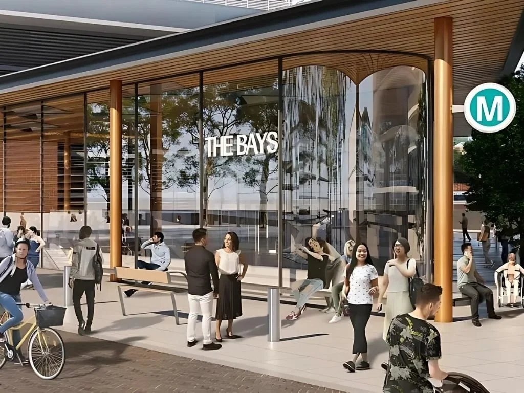 Rendering of a modern bus stop with people waiting and socializing, surrounded by trees, with a glass shelter displaying 'THE BAYS' sign and an 'M' transit marker.