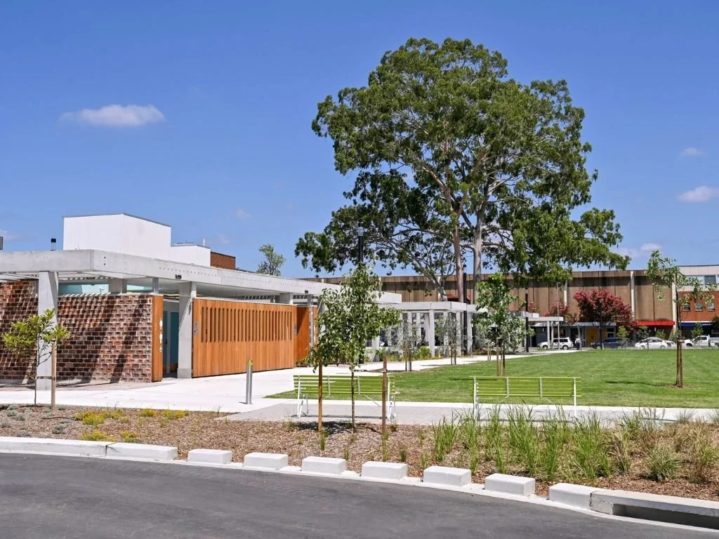A modern park or public space with a large tree, green grass, benches, and a pathway, adjacent to a building with a white and brown exterior.