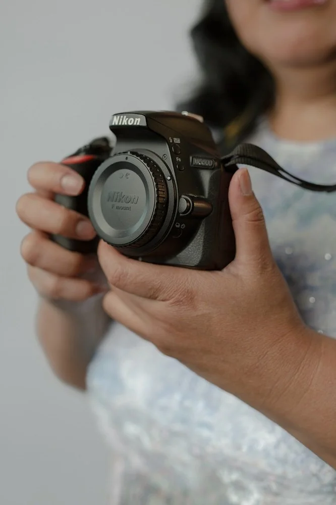 A person holding a black Nikon camera with a lens cap on, with a gray background.