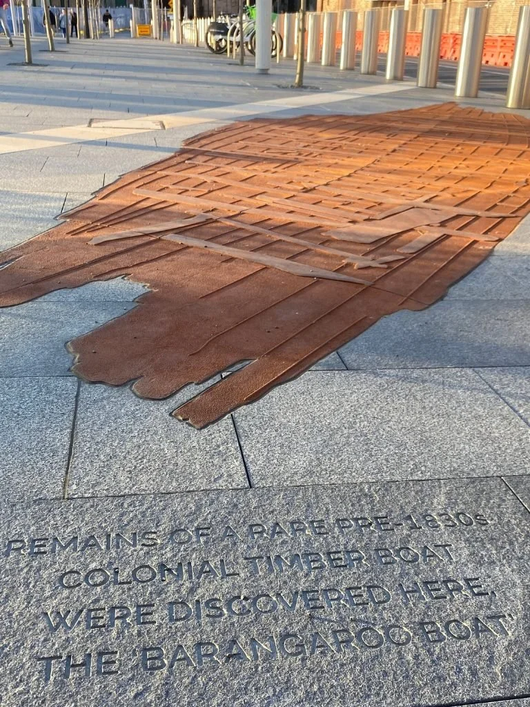 Remains of a colonial timber boat displayed on the ground with an inscription about its discovery, on a paved outdoor area with a fence, poles, and parked bikes in the background.
