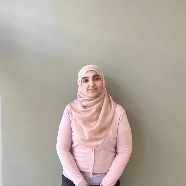 A woman wearing a pink headscarf and light pink cardigan standing against a plain beige wall.