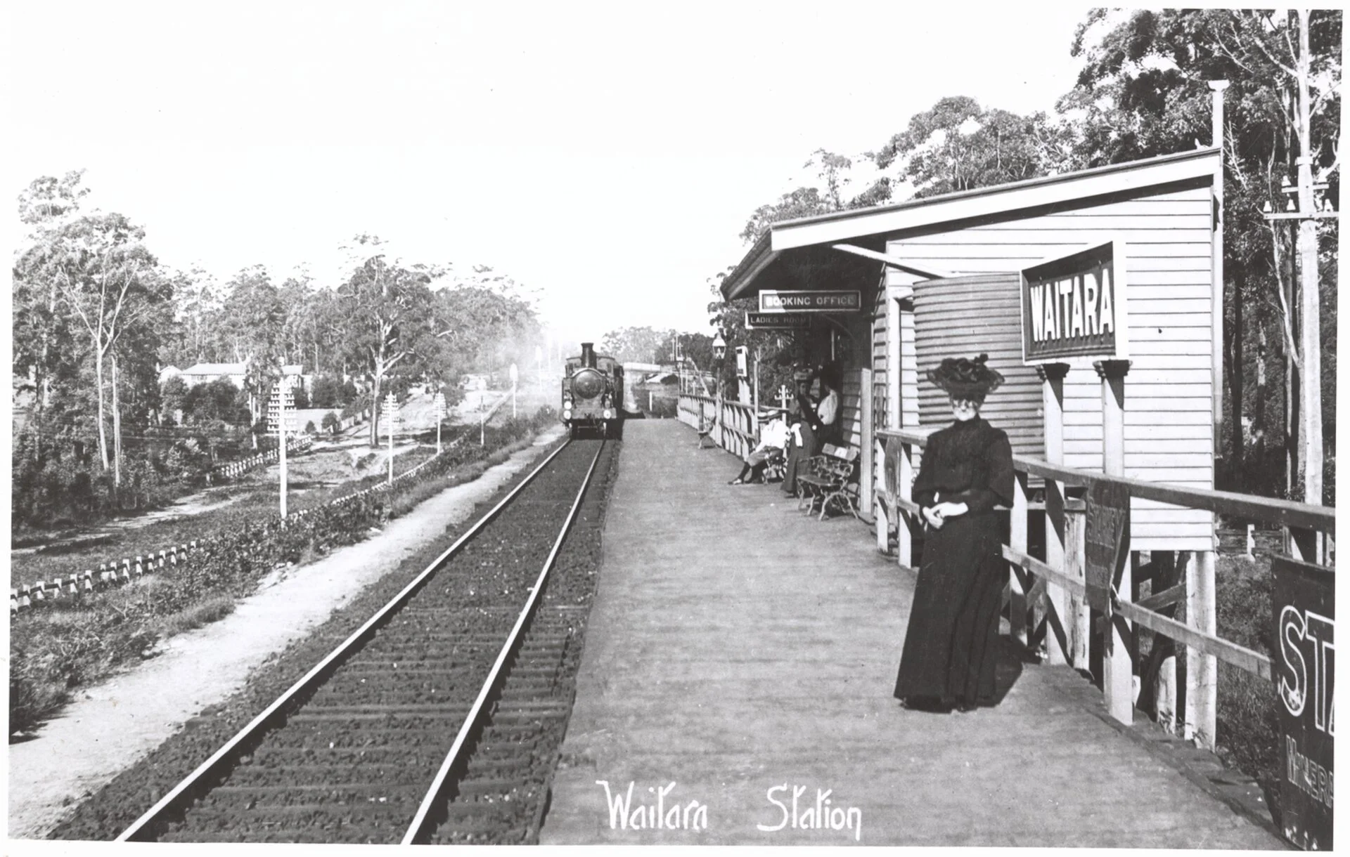 A black-and-white photo of Wailara Station with a train approaching, several people waiting on the platform, a booking office, and a building labeled WAILARA.