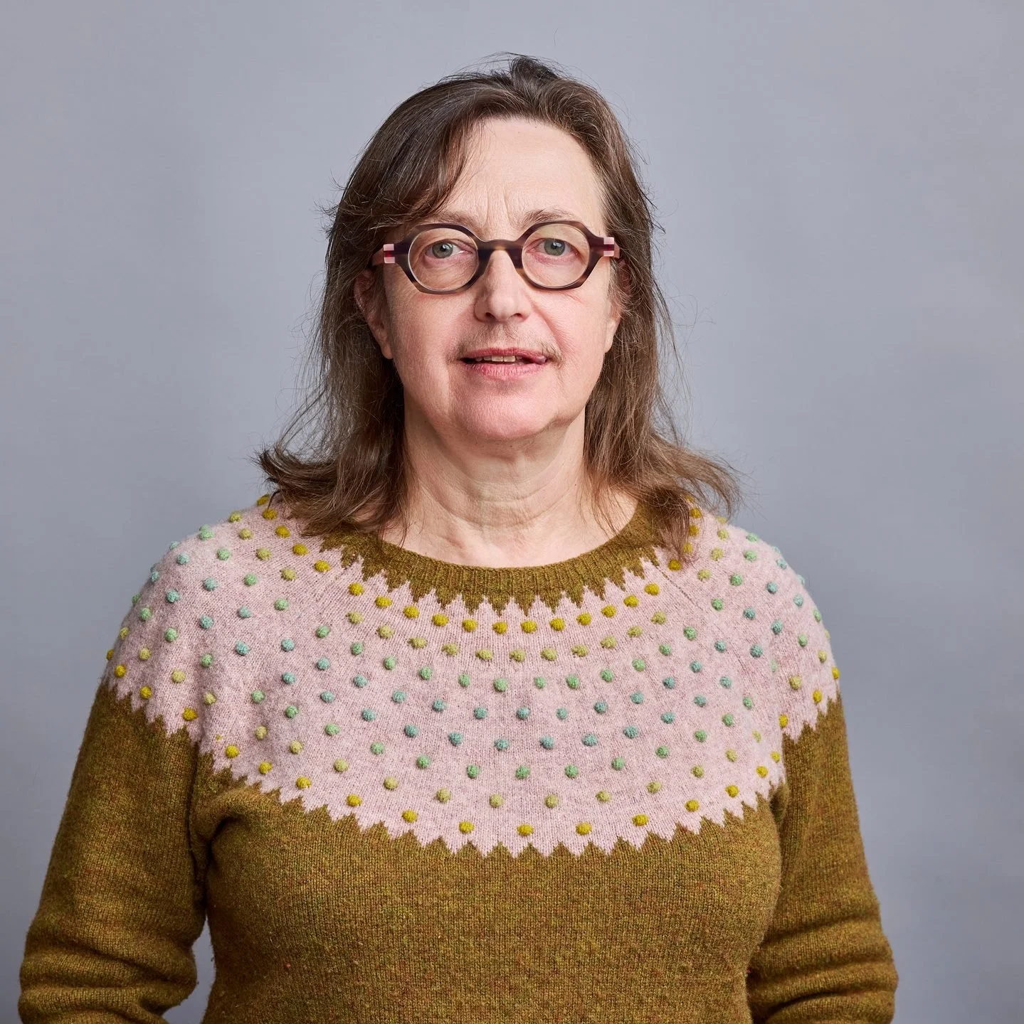 A middle-aged woman with shoulder-length brown hair and glasses, wearing a pink and brown knitted sweater with a dotted pattern, standing against a plain gray background.