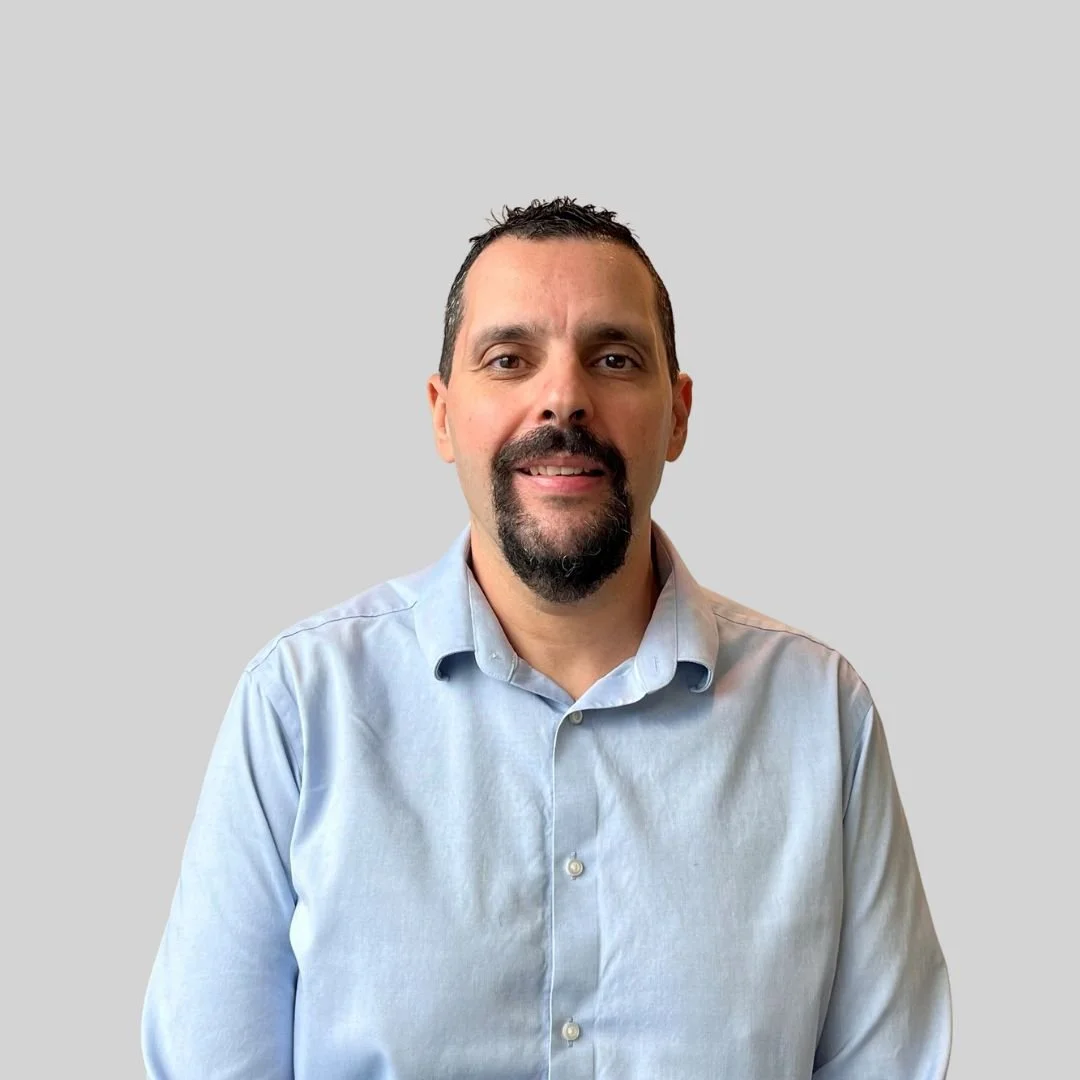 A man with dark hair and a beard wearing a light blue button-up shirt facing the camera against a plain gray background.