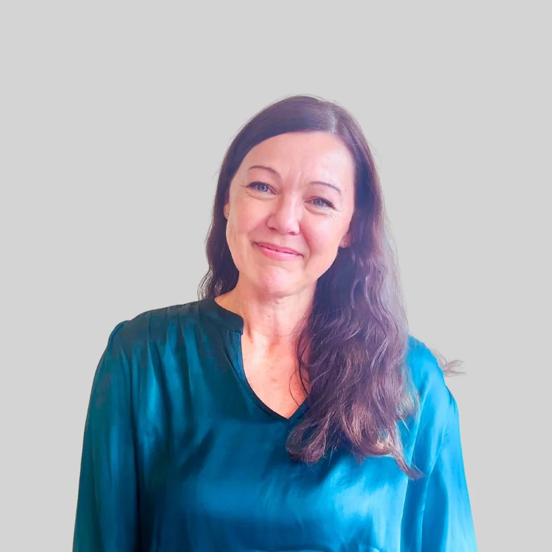 A woman with long wavy brown hair wearing a blue satin blouse, smiling softly, with a plain light gray background.