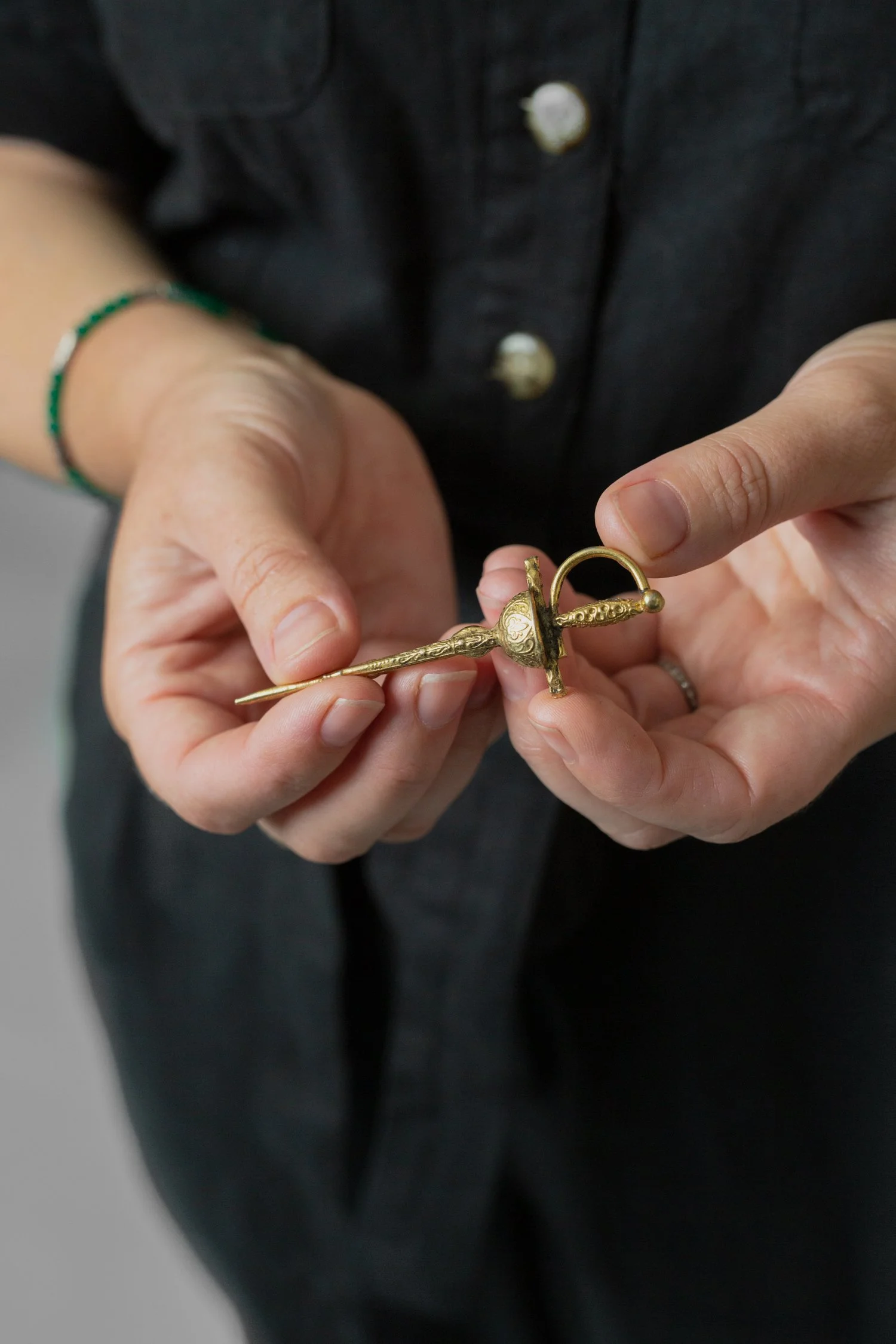 Close-up of a person holding a small, ornate, gold-colored sword or dagger with a rounded handle and detailed blade, wearing a dark shirt with pearl buttons and a green bracelet on their wrist.