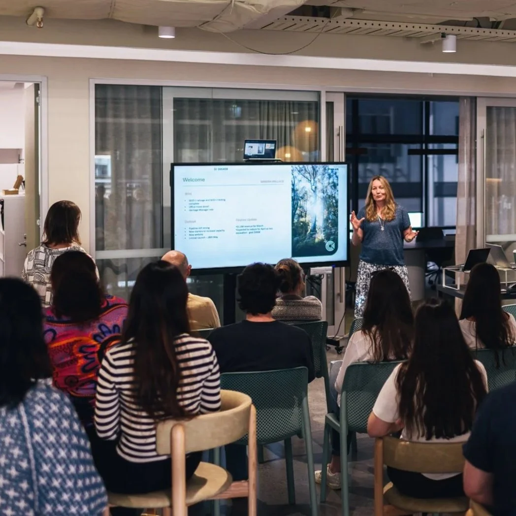 A woman giving a presentation to a group of people in a conference room with a large digital screen displaying a slide titled 'Welcome' and a nature scene on the right side.