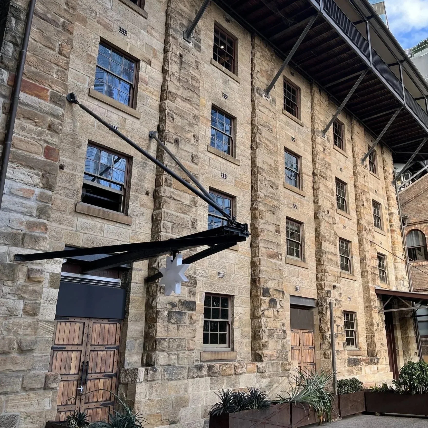 A stone building with multiple windows and wooden doors. An outdoor balcony and a hanging star decoration are visible, along with some plants at the base.