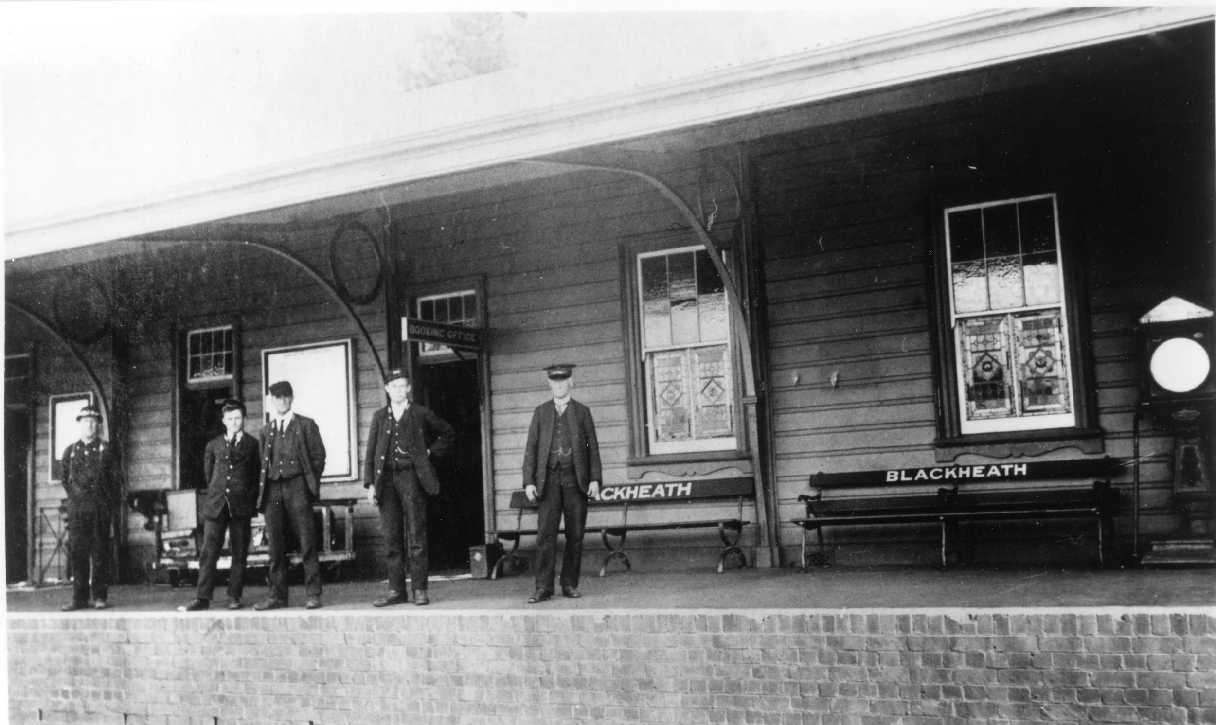Black and white photo of a train station platform with four uniformed personnel standing in front of benches labeled 'Blackheath'. There are three windows with decorative stained glass and a small sign that reads 'Booking Office'.