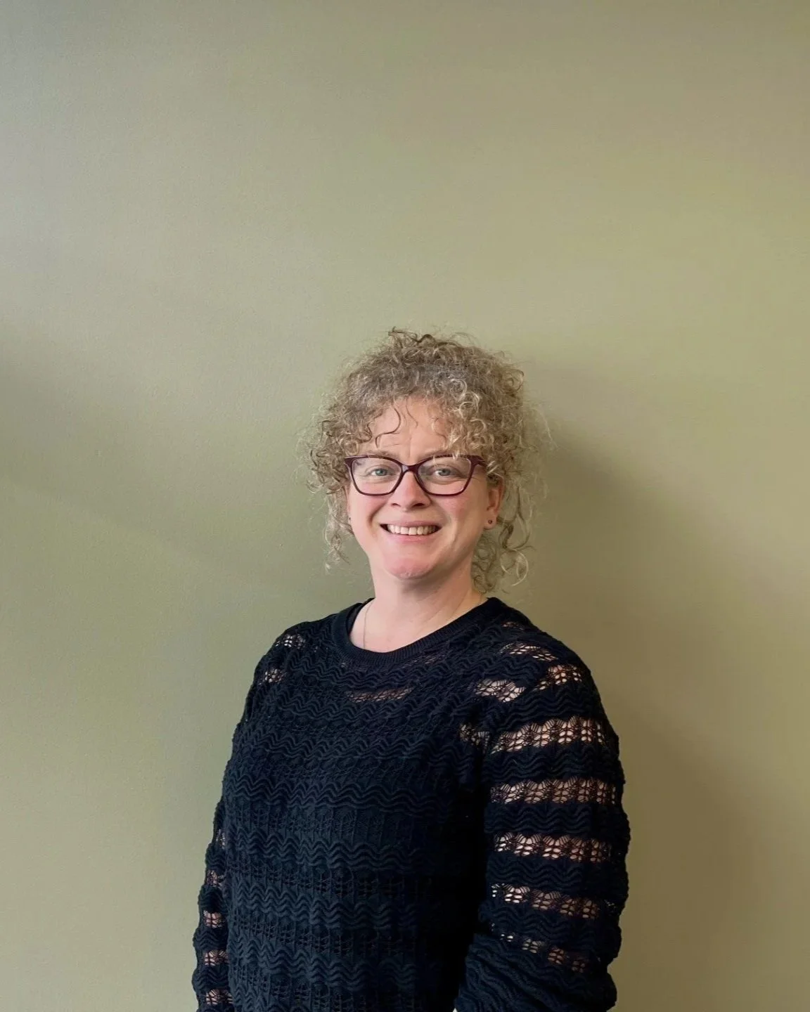 A woman with curly blonde hair, glasses, and a black lace top standing against a beige wall, smiling