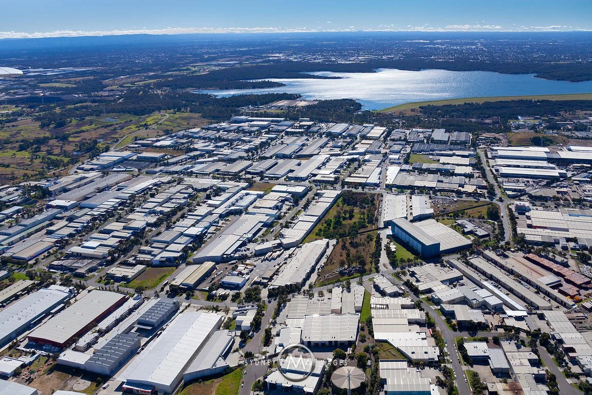 Aerial view of an industrial park with numerous warehouses and buildings, surrounding a lake with greenery and open land in the background.