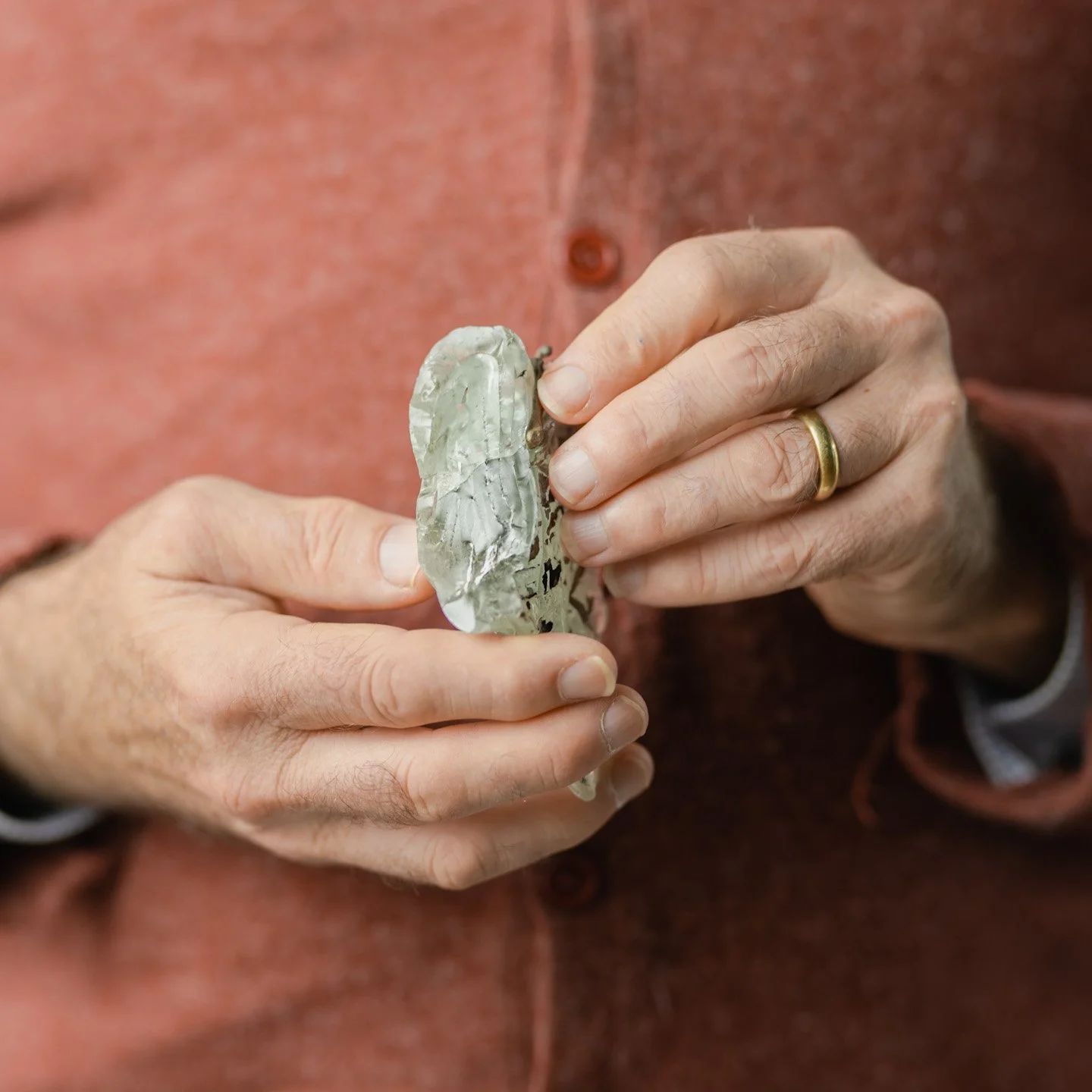 Close-up of a person holding a large, irregularly shaped piece of mother-of-pearl shell with both hands, wearing a gold ring on their finger, dressed in a reddish-brown shirt.