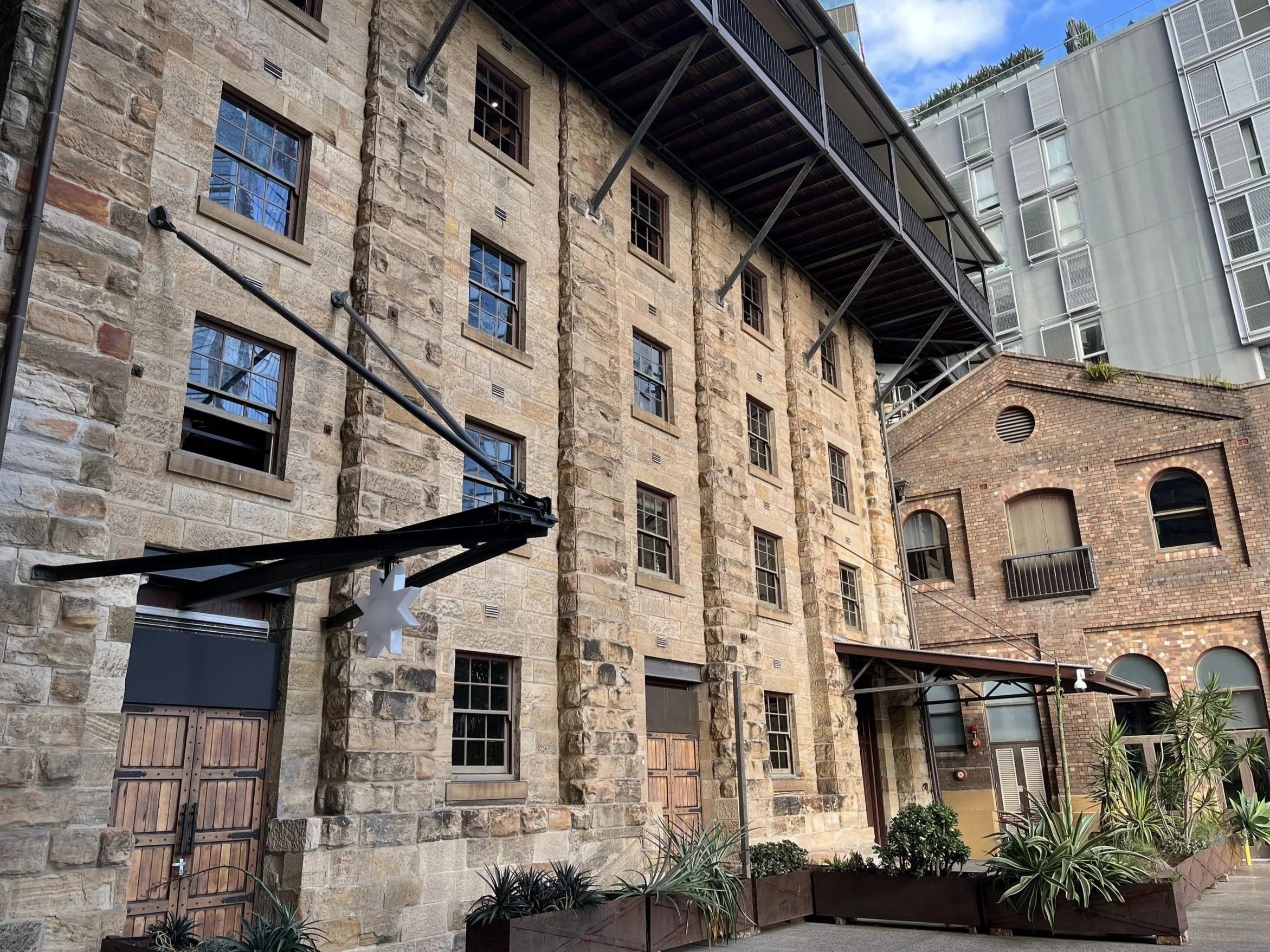 View of multi-story buildings with brick and stone facades, metal fire escapes, and surrounding plants, under a partly cloudy sky.