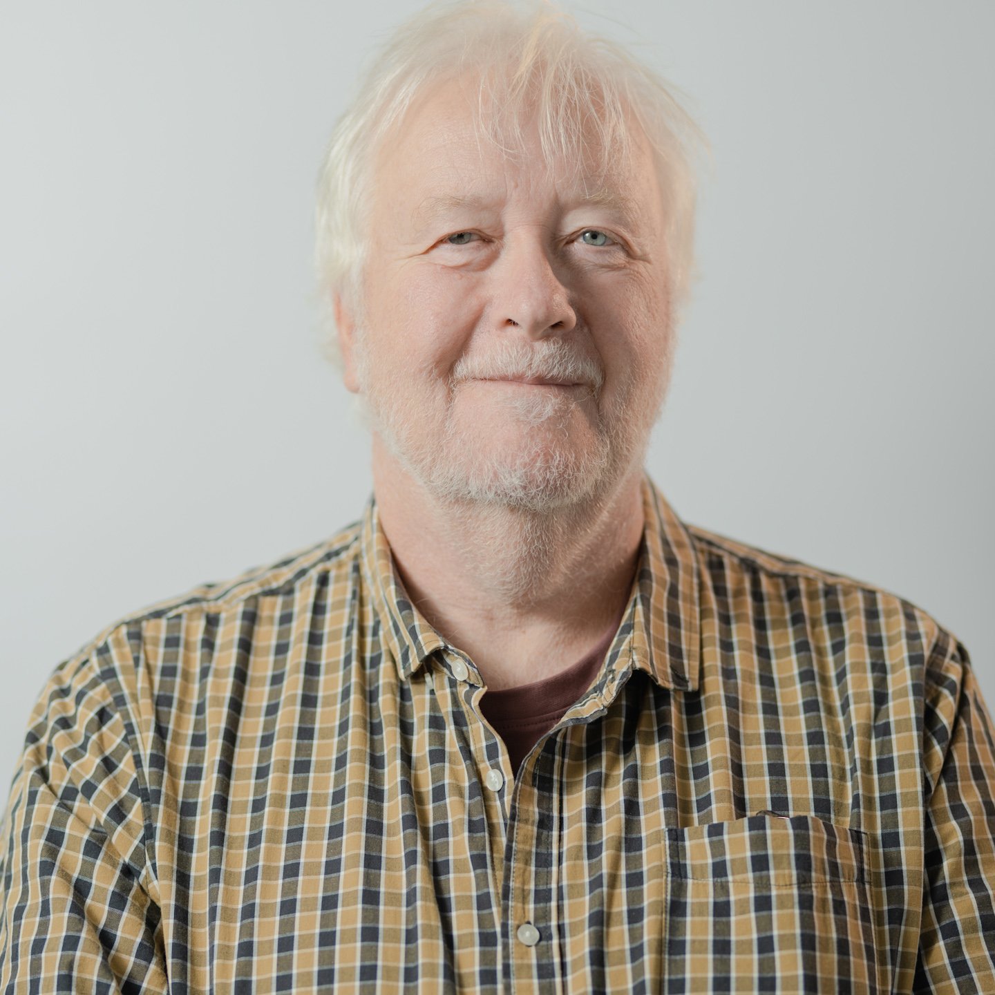 A middle-aged man with gray hair and a beard, smiling slightly, wearing a checkered shirt against a plain light gray background.