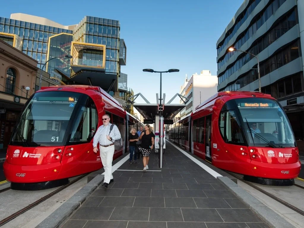 Two red modern light rail trains at a city station with pedestrians walking between them, surrounded by tall buildings.