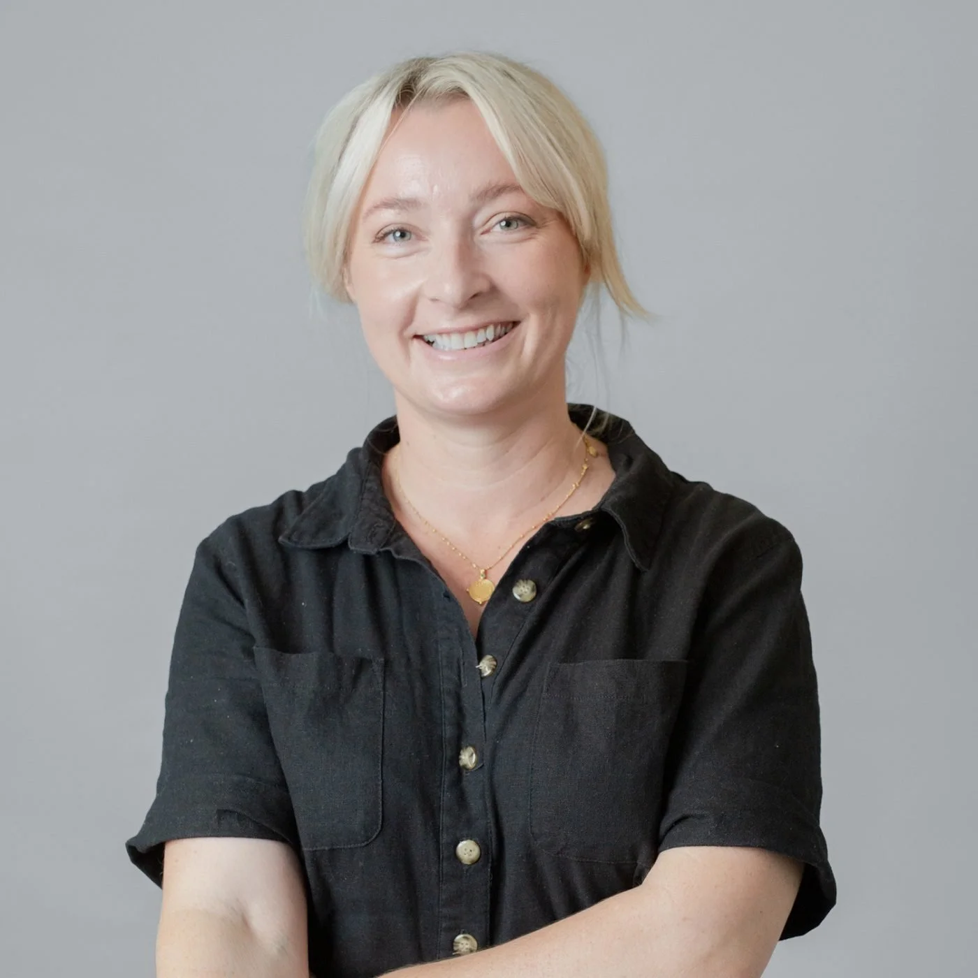 A smiling woman with short blonde hair, wearing a black button-up shirt and a gold necklace, standing against a plain gray background.