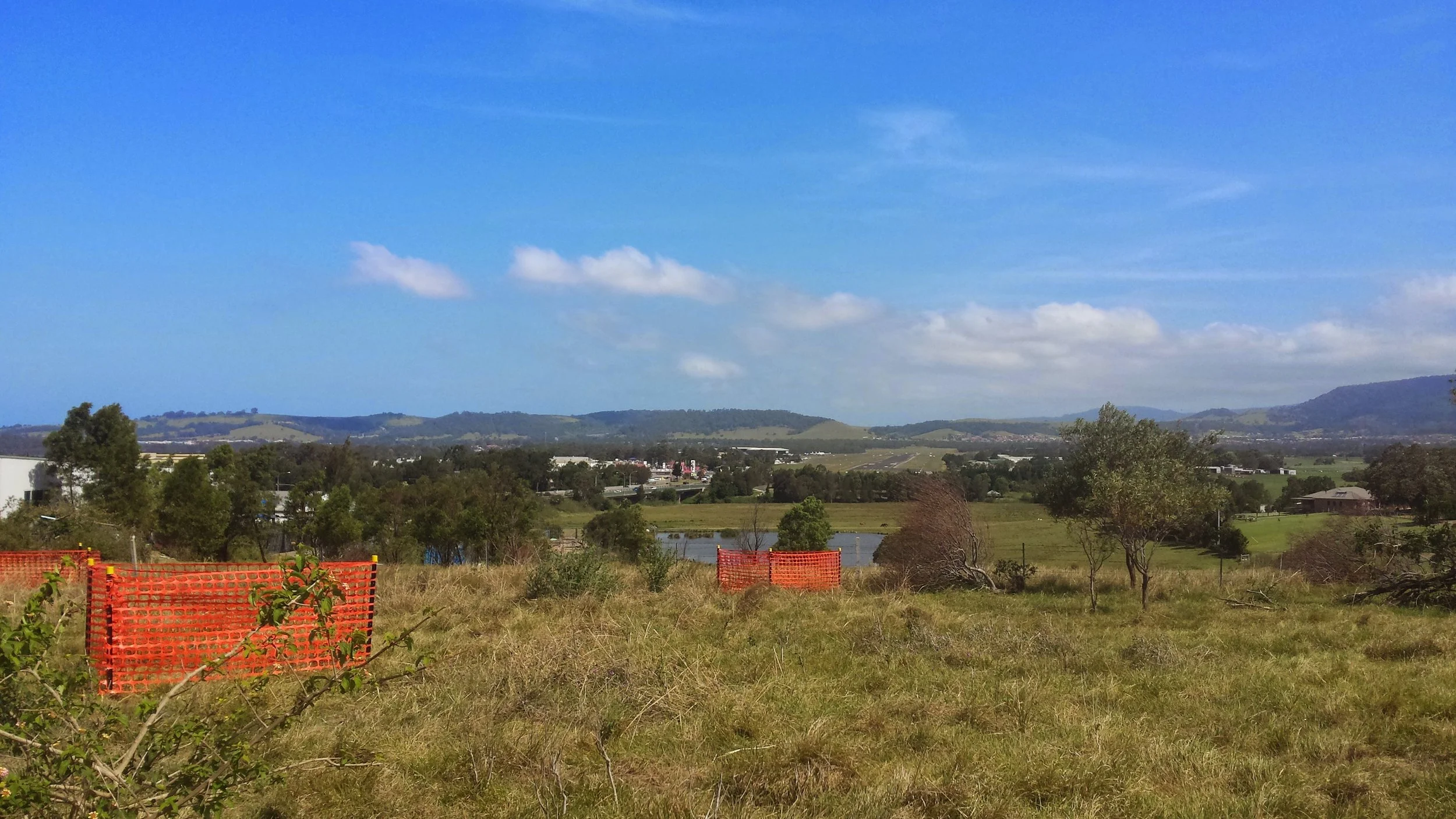 Landscape view of a rural area with trees, a small pond, and rolling hills under a blue sky with scattered clouds.