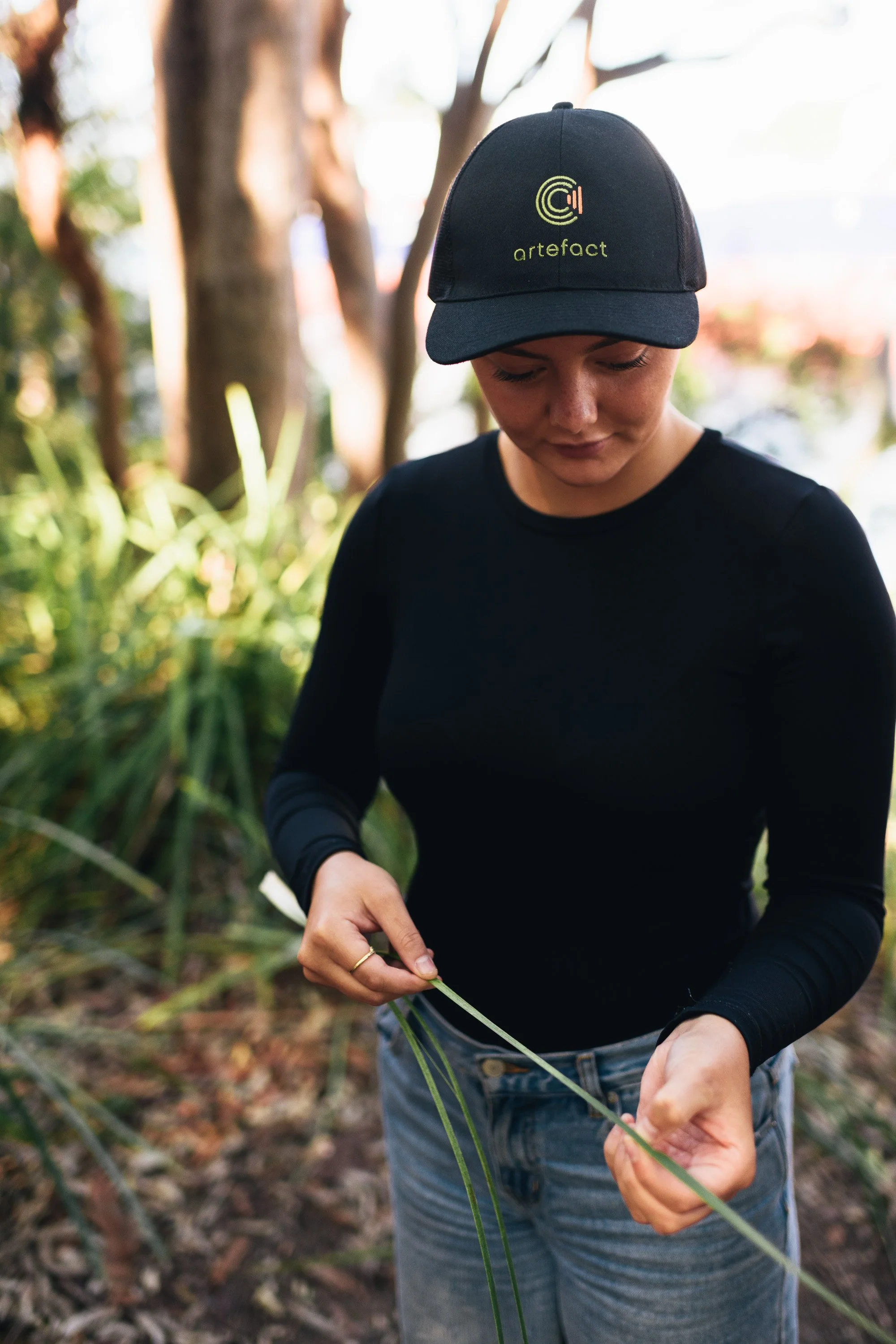 A woman with a black cap and black long-sleeve shirt is outdoors, handling green grass or plant stems with both hands. There are trees and greenery in the background.