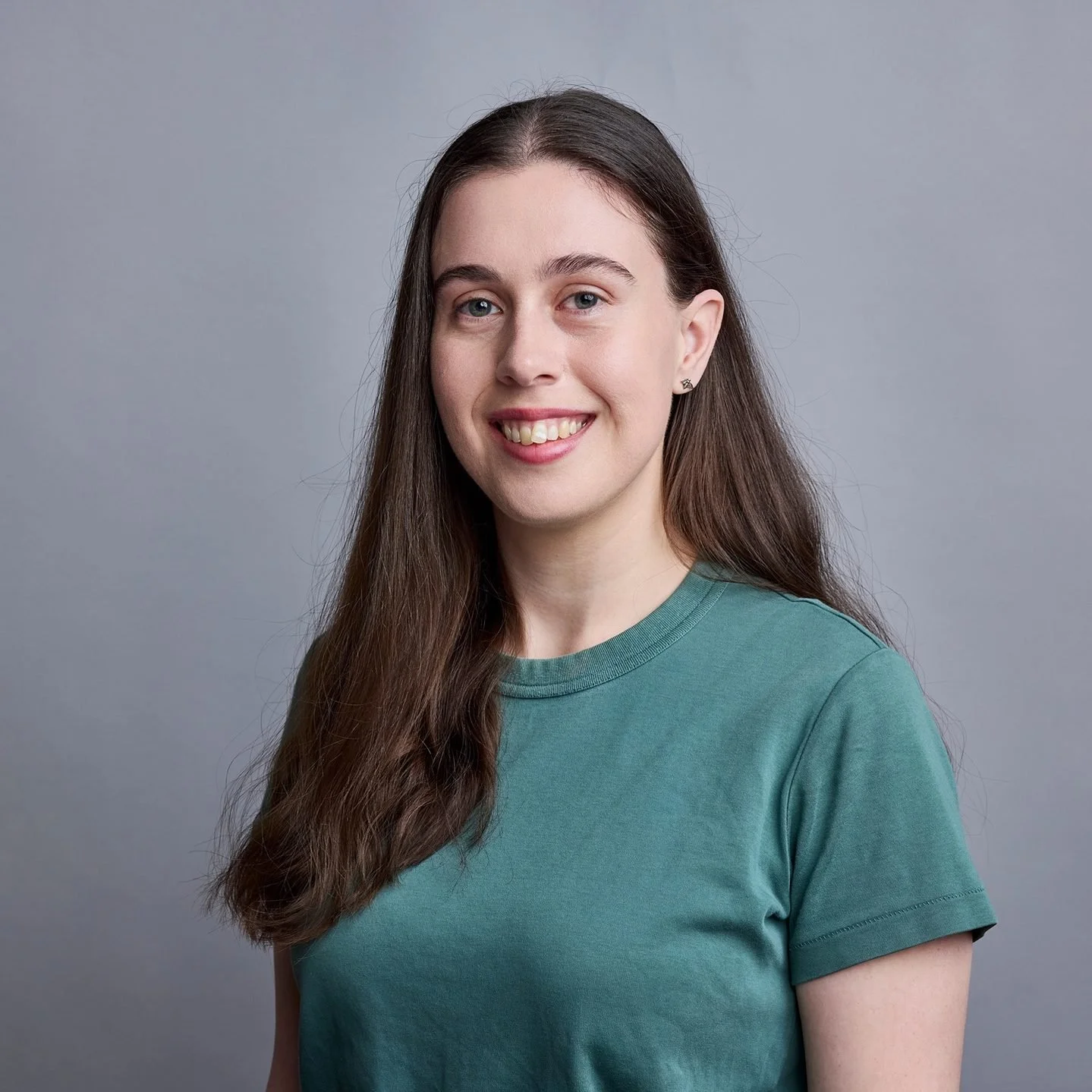 Portrait of a young woman with long brown hair, wearing a green T-shirt, smiling, against a gray background.