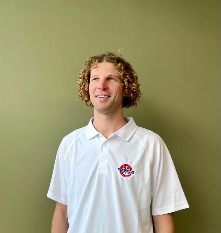 A man with curly blond hair smiling, wearing a white polo shirt with a logo on the chest, standing against a light green wall.