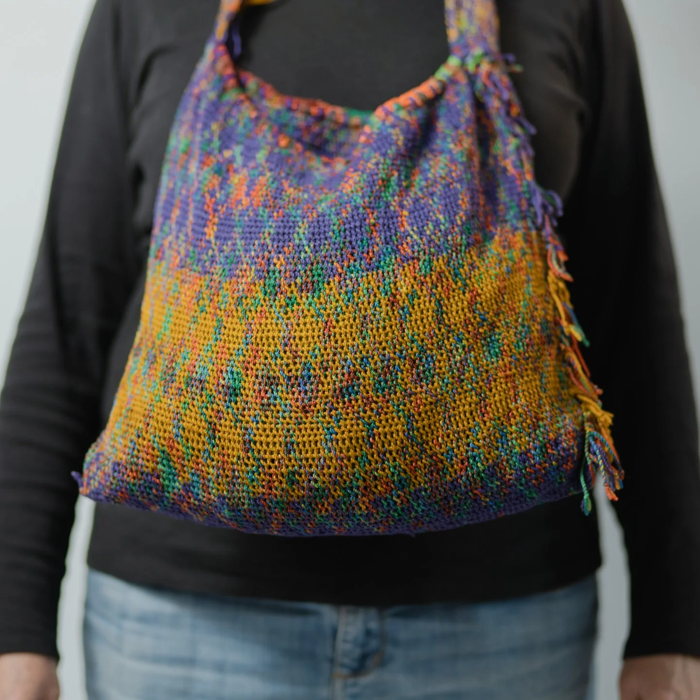 Person wearing a black long-sleeve shirt and blue jeans, with a multicolored woven bag on their shoulder.