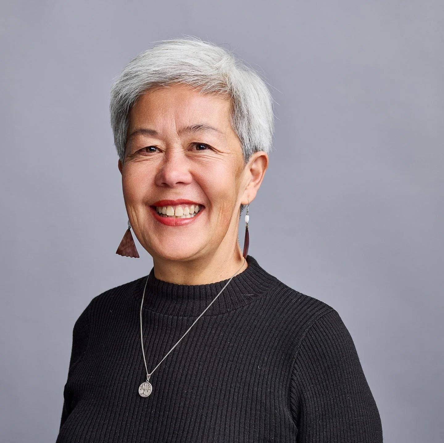 A smiling woman with short gray hair, wearing a black ribbed top, earrings, and a necklace, against a gray background.
