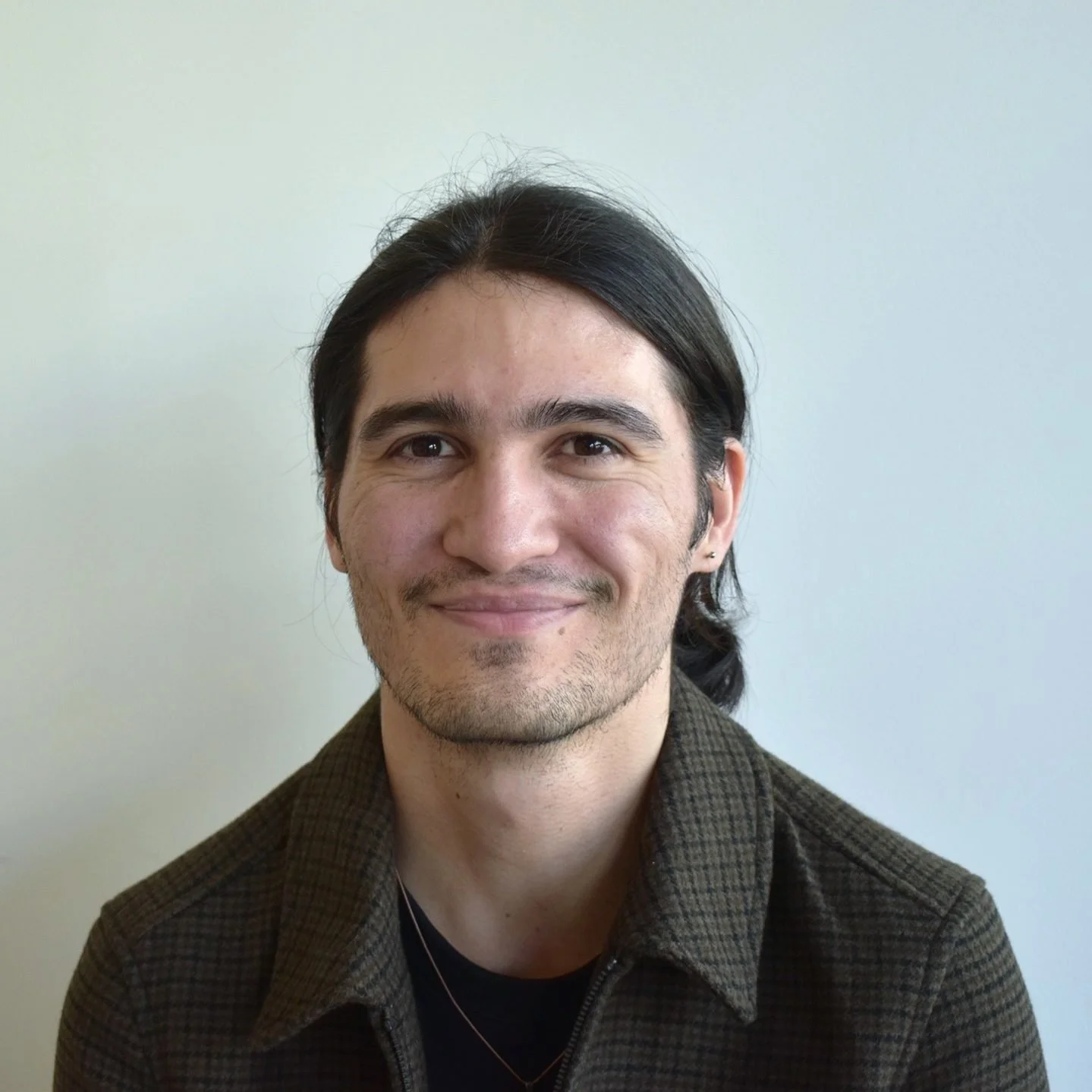 A man with dark hair, facial hair, and light skin, smiling while facing the camera, wearing a dark shirt and a plaid brown jacket, standing against a plain light-colored background.